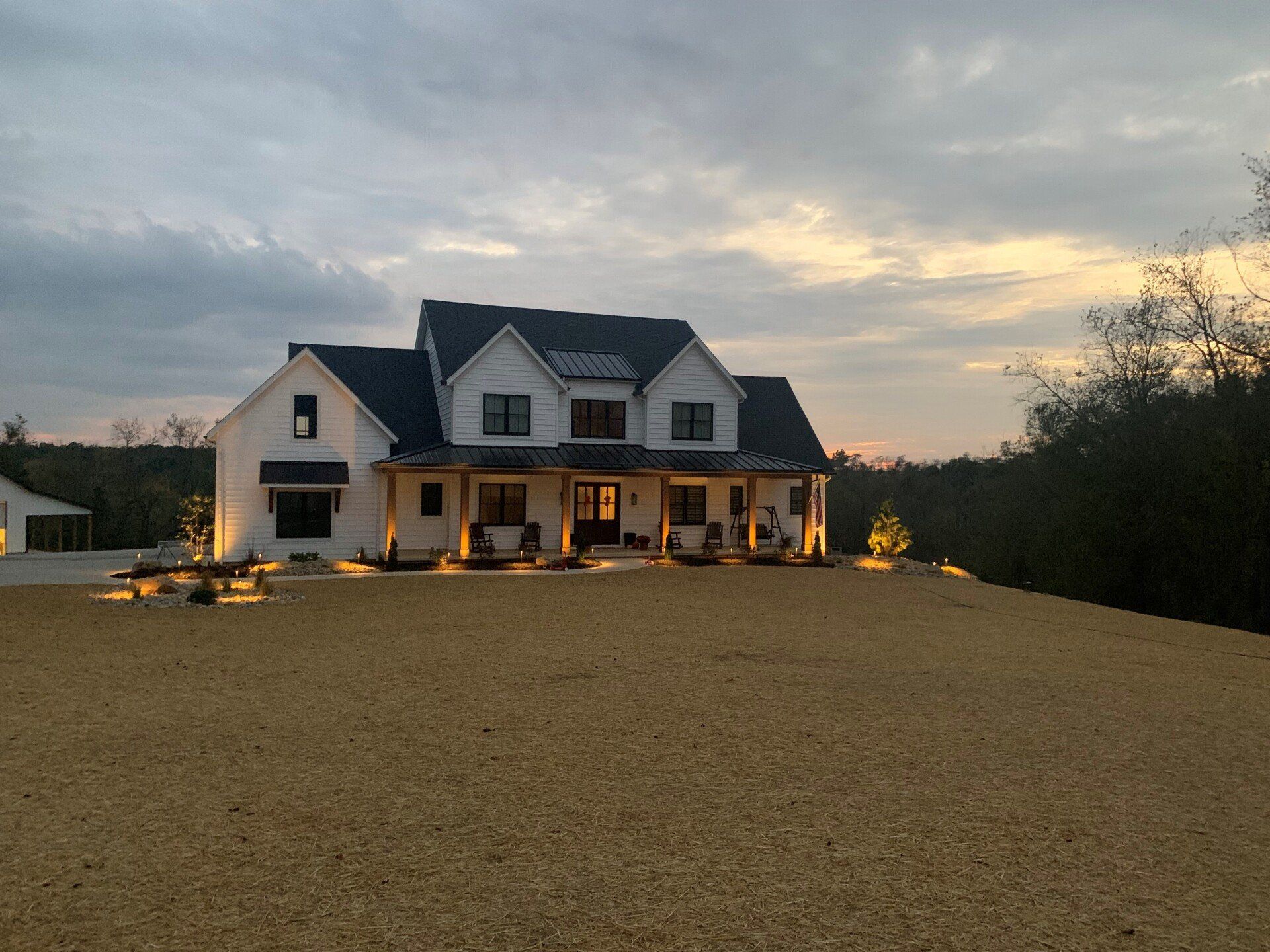 A white two-story modern farmhouse with a dark roof and warm exterior lighting at twilight, set against a vast field.