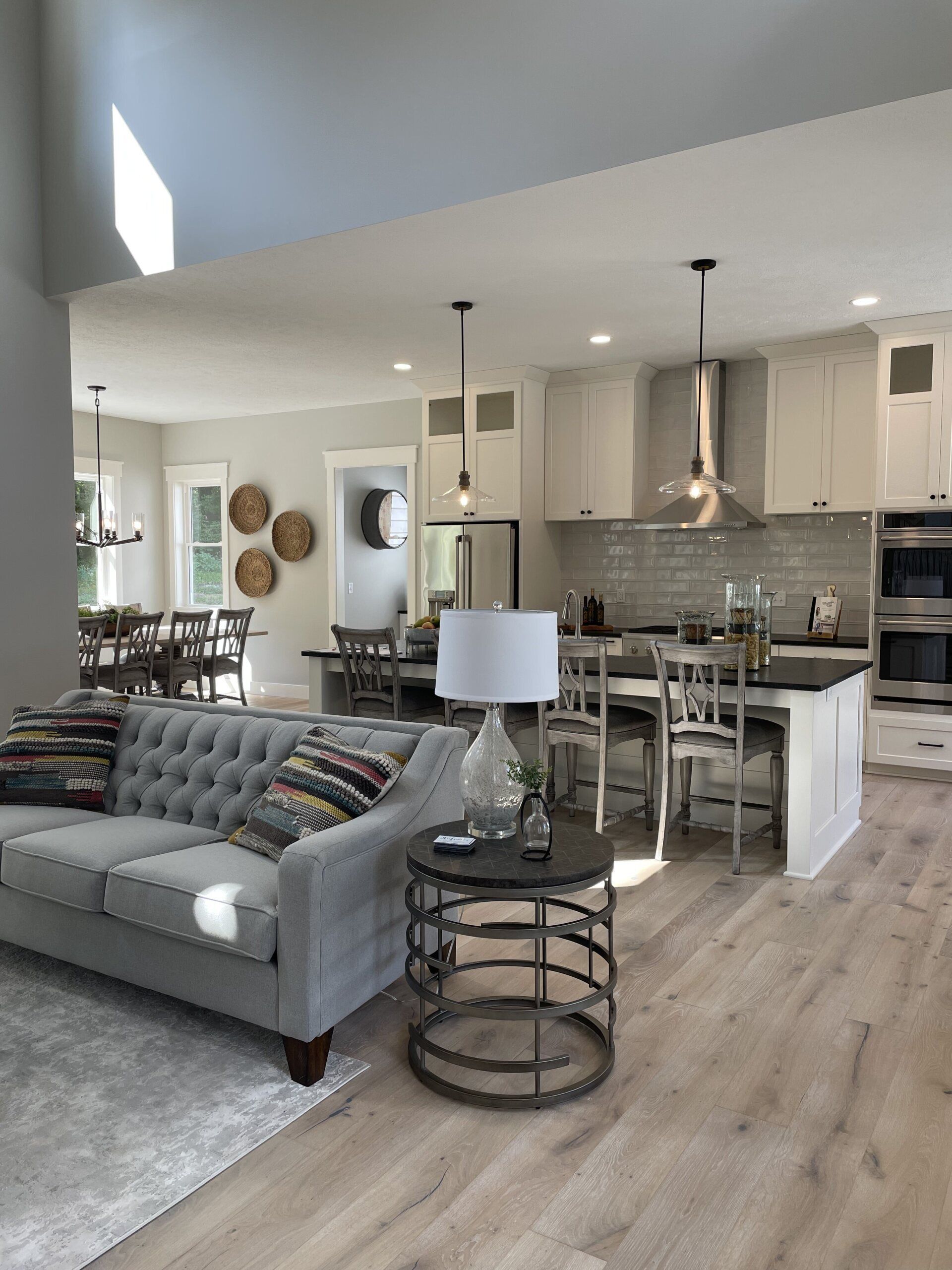 A bright, open-concept living room and kitchen with a grey tufted sofa, round metal end table, and kitchen island seating.