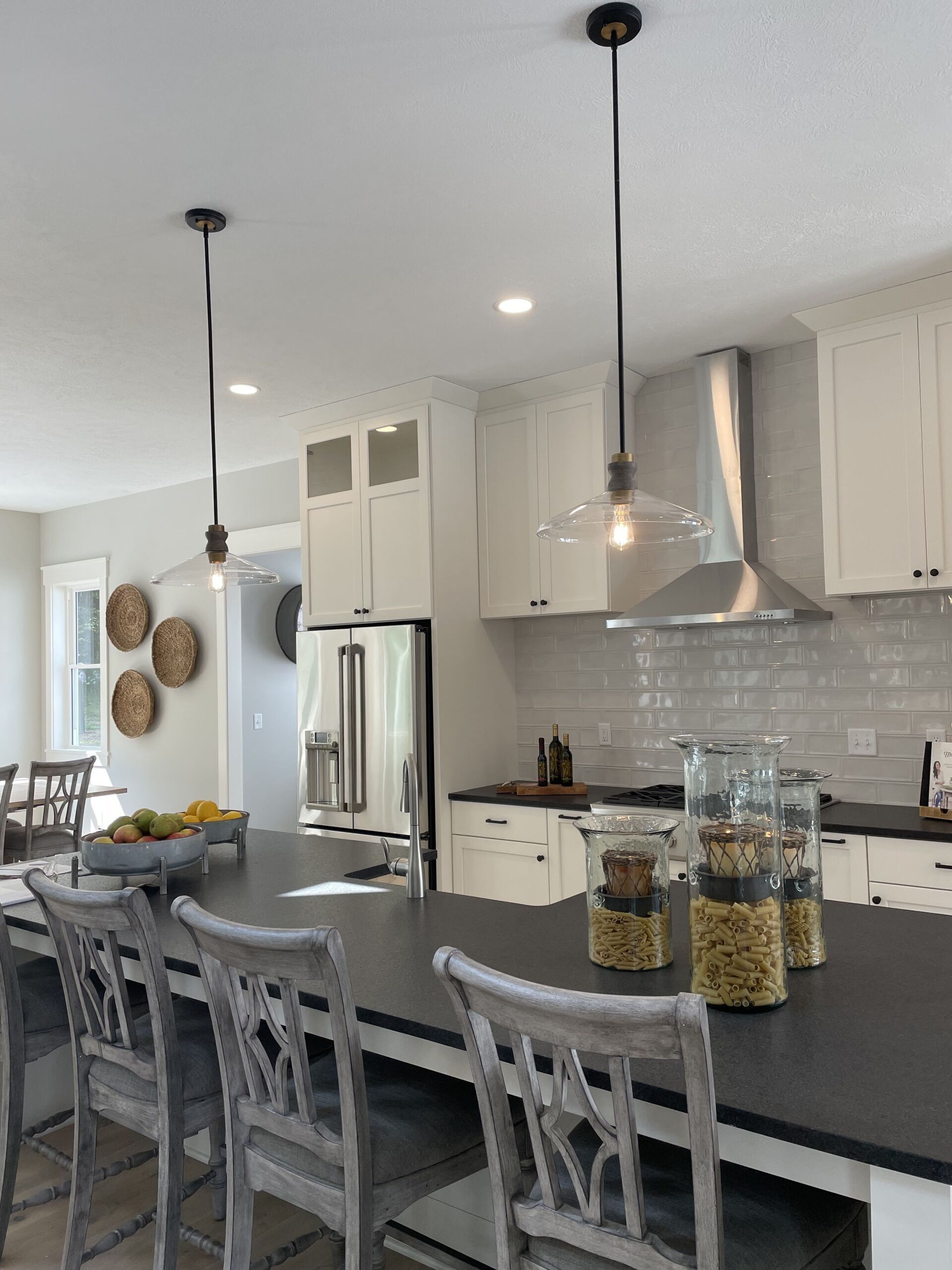A kitchen with white cabinets, a dark island, gray chairs, two hanging pendant lights, and a stainless steel range hood.