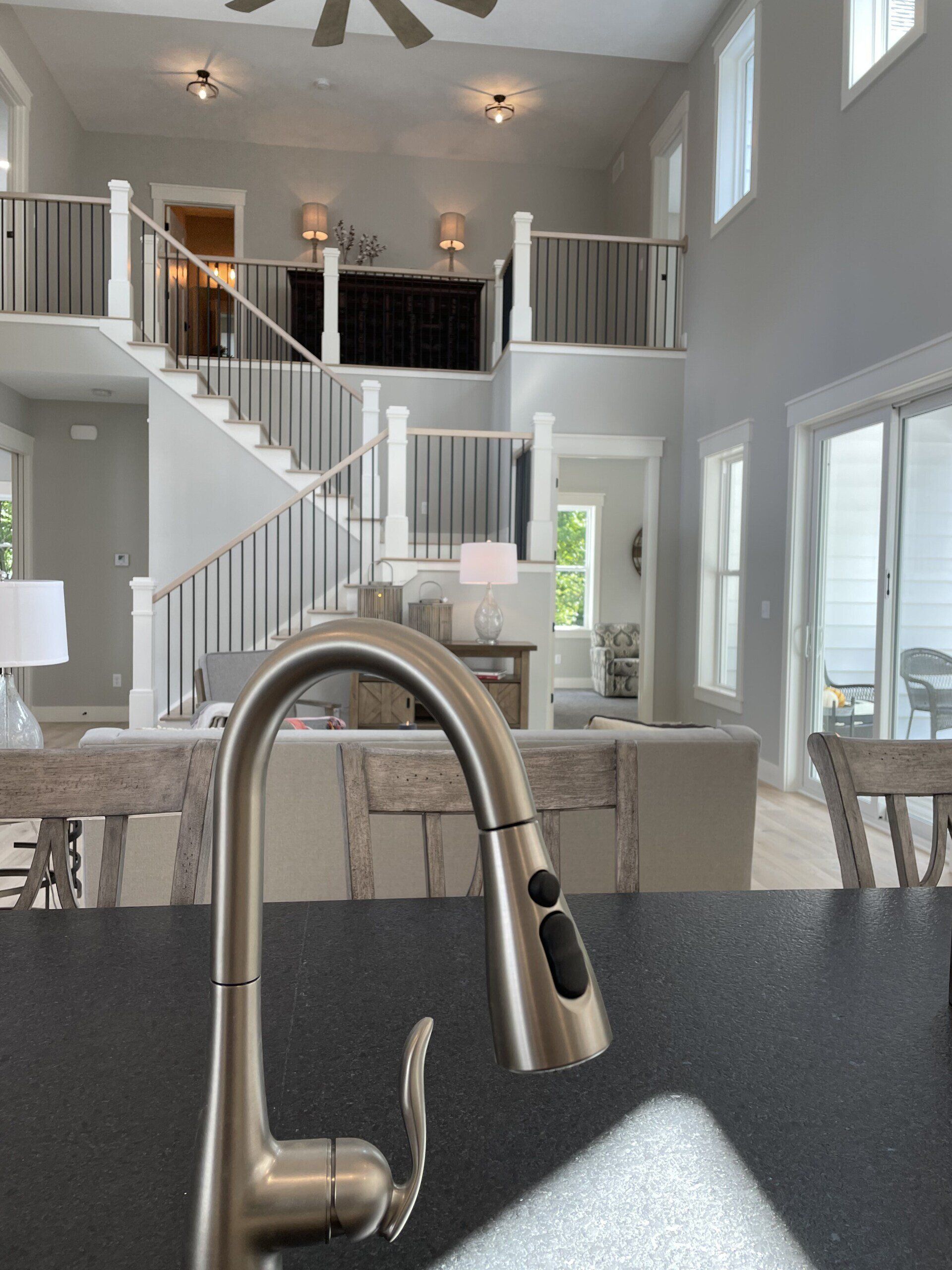 A metallic kitchen faucet in the foreground with an out-of-focus view of a bright, open-concept living area and staircase.