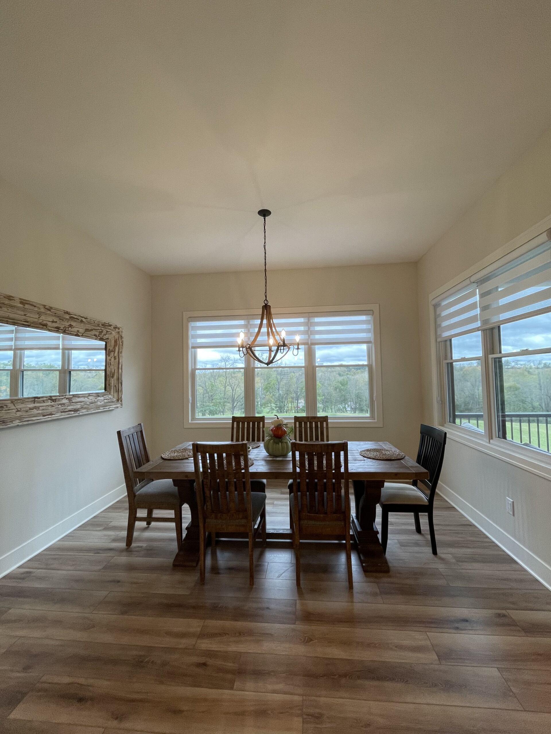 A dining room with a wooden table, six chairs, a chandelier, and a mirror on the wall, overlooking a view of trees.