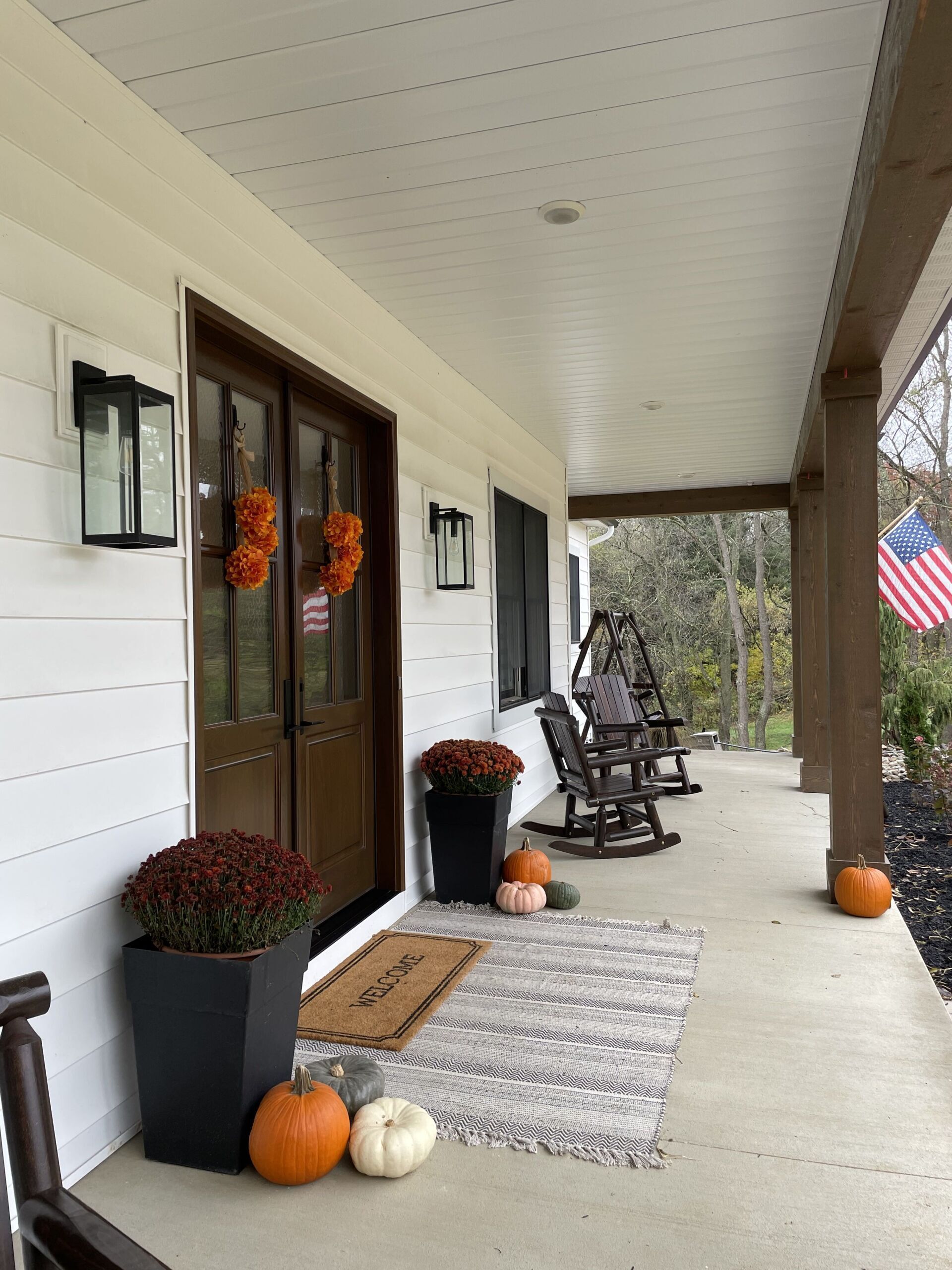 A covered front porch decorated for autumn with pumpkins, potted mums, rocking chairs, and an American flag.