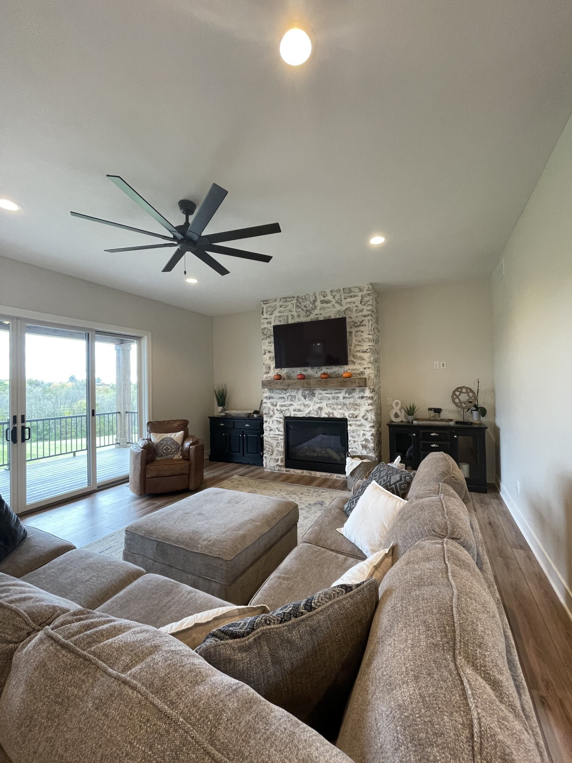 A living room with a stone fireplace, dark ceiling fan, large gray sectional sofa, ottoman, and sliding glass balcony door.