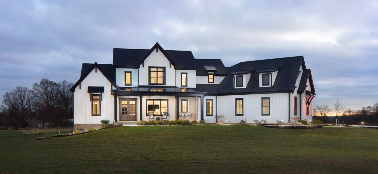 A modern two-story farmhouse with a white exterior, dark roof, and multiple gables, set against a lawn at dusk.