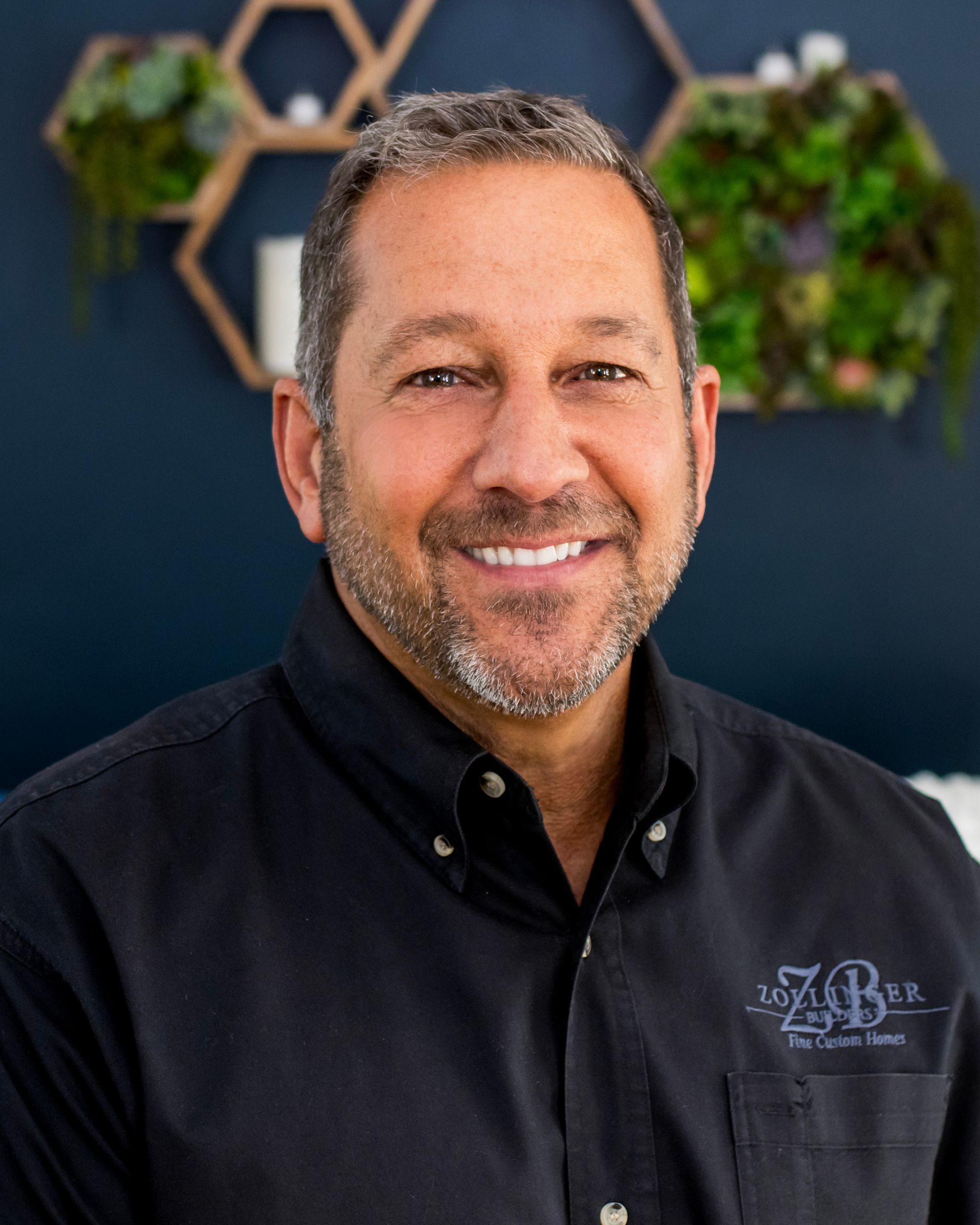 A smiling person wearing a black button-down shirt stands in front of a dark blue wall with decorative hexagonal shelves.