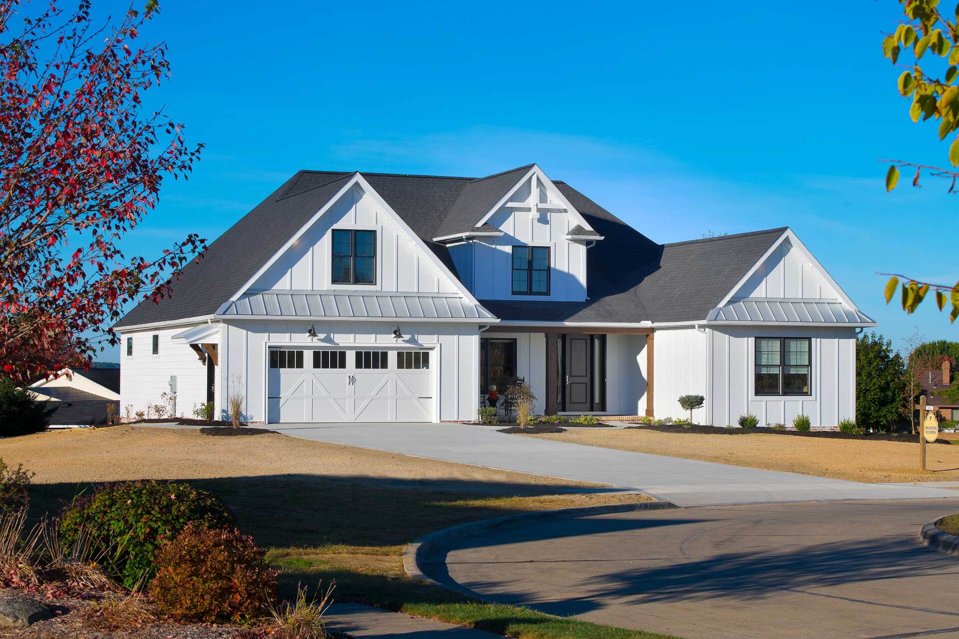 A modern white farmhouse with a gray roof and a two-car garage, surrounded by a yard with dry grass under a blue sky.
