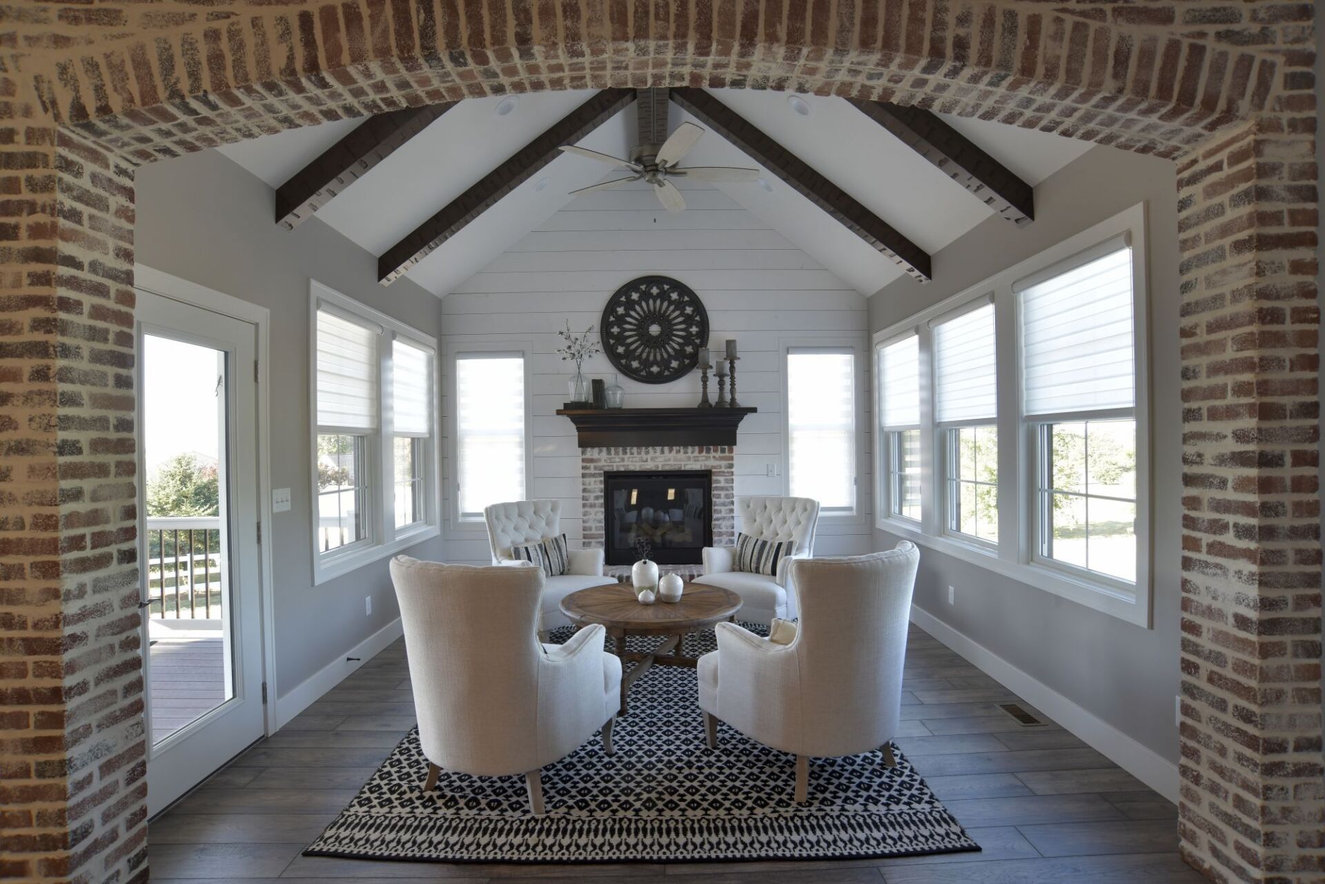 A sunroom with a brick-arched entry, two cream chairs, a central coffee table on a patterned rug, and a fireplace.