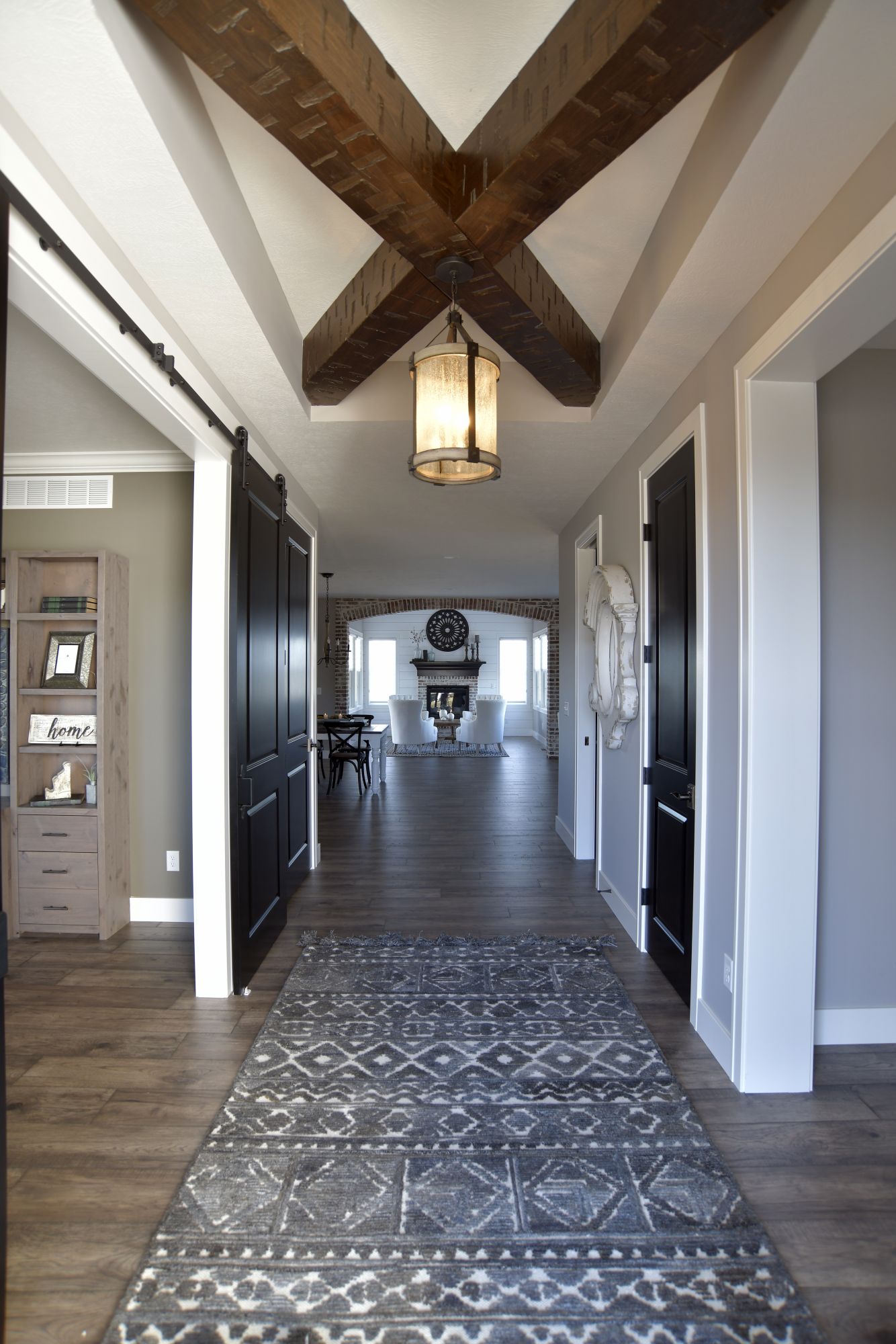 A view down a hallway with an X-patterned wood beam ceiling, black doors, and a patterned rug leading to a living room.