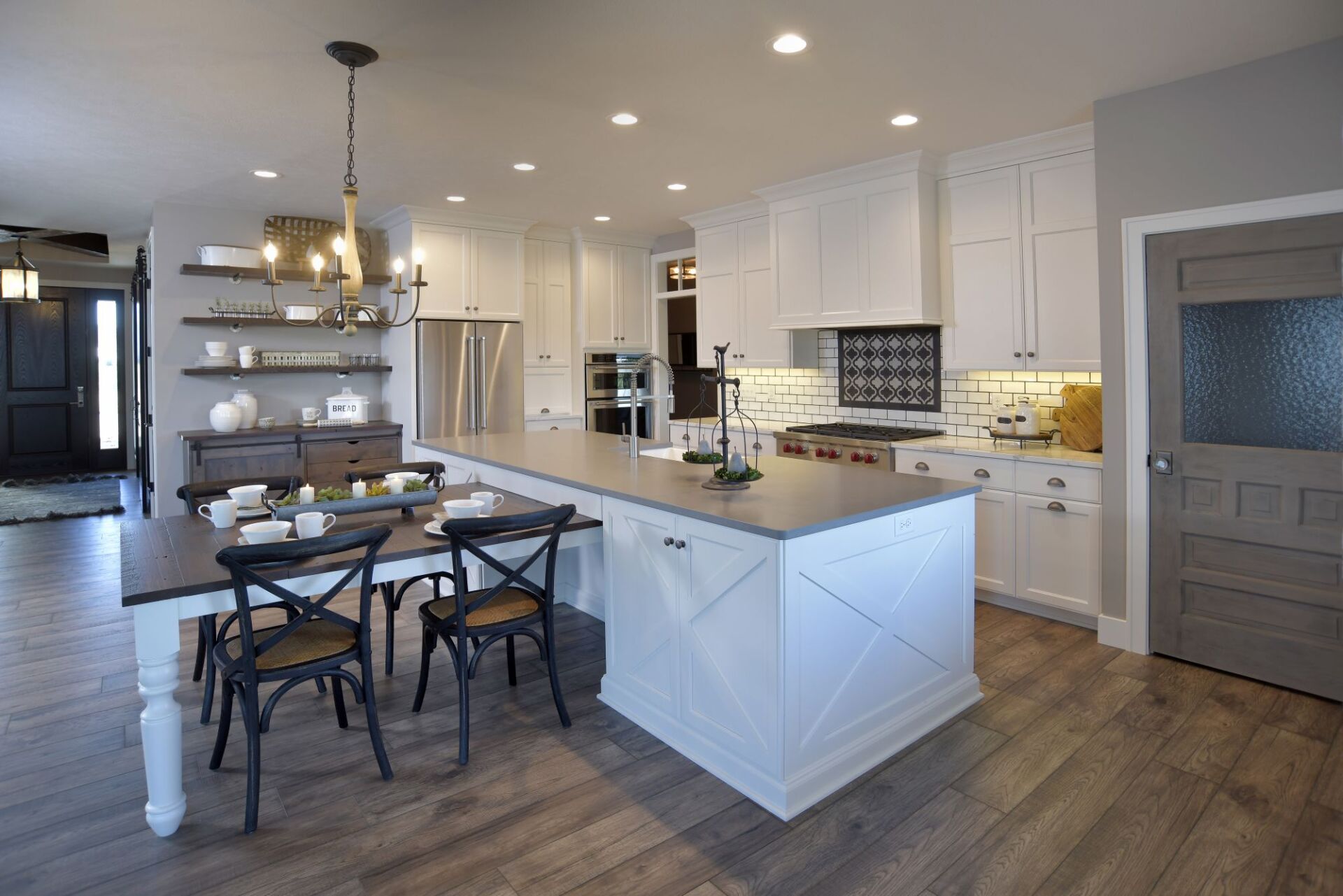 A modern farmhouse kitchen with white cabinets, a large island with a table extension, and light wood-plank flooring.