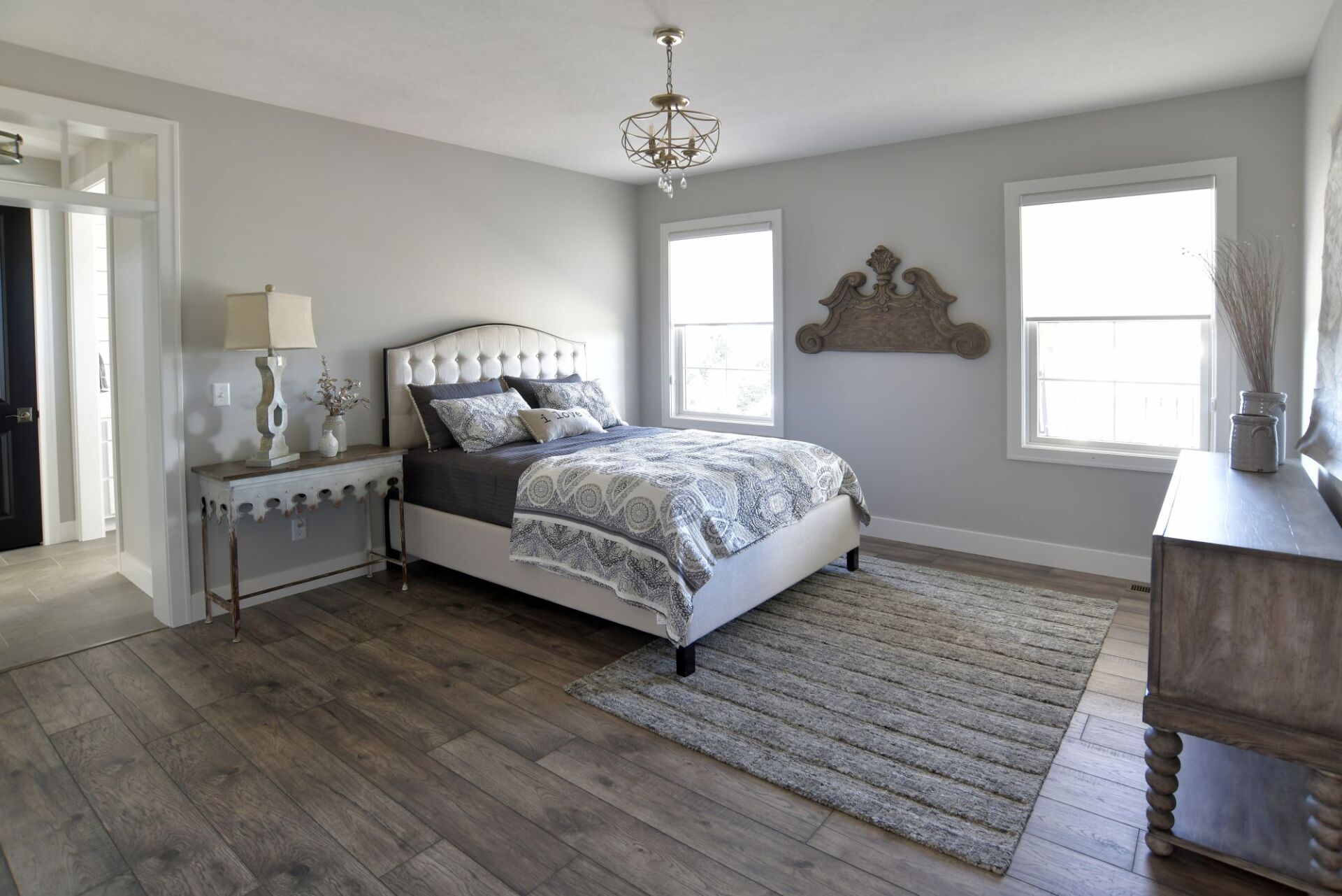 A modern bedroom with a tufted bed, grey bedding, wooden floors, area rug, and wooden furniture against light grey walls.