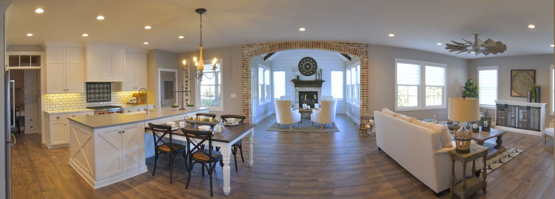 A wide-angle interior view of an open-concept kitchen and living area with white cabinets, wood floors, and a fireplace.