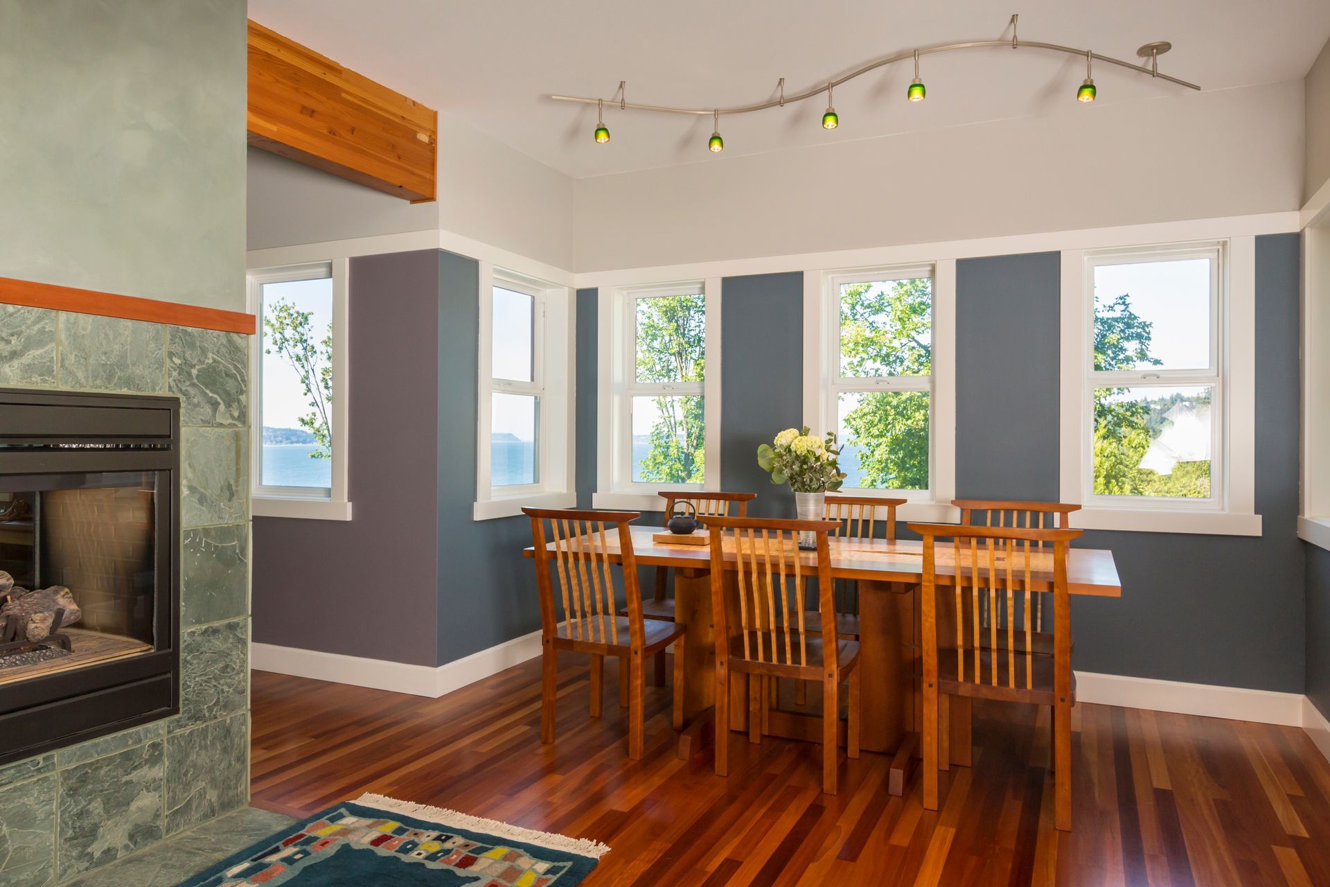 Dining room with wooden table, chairs, large windows, and a fireplace on the left.