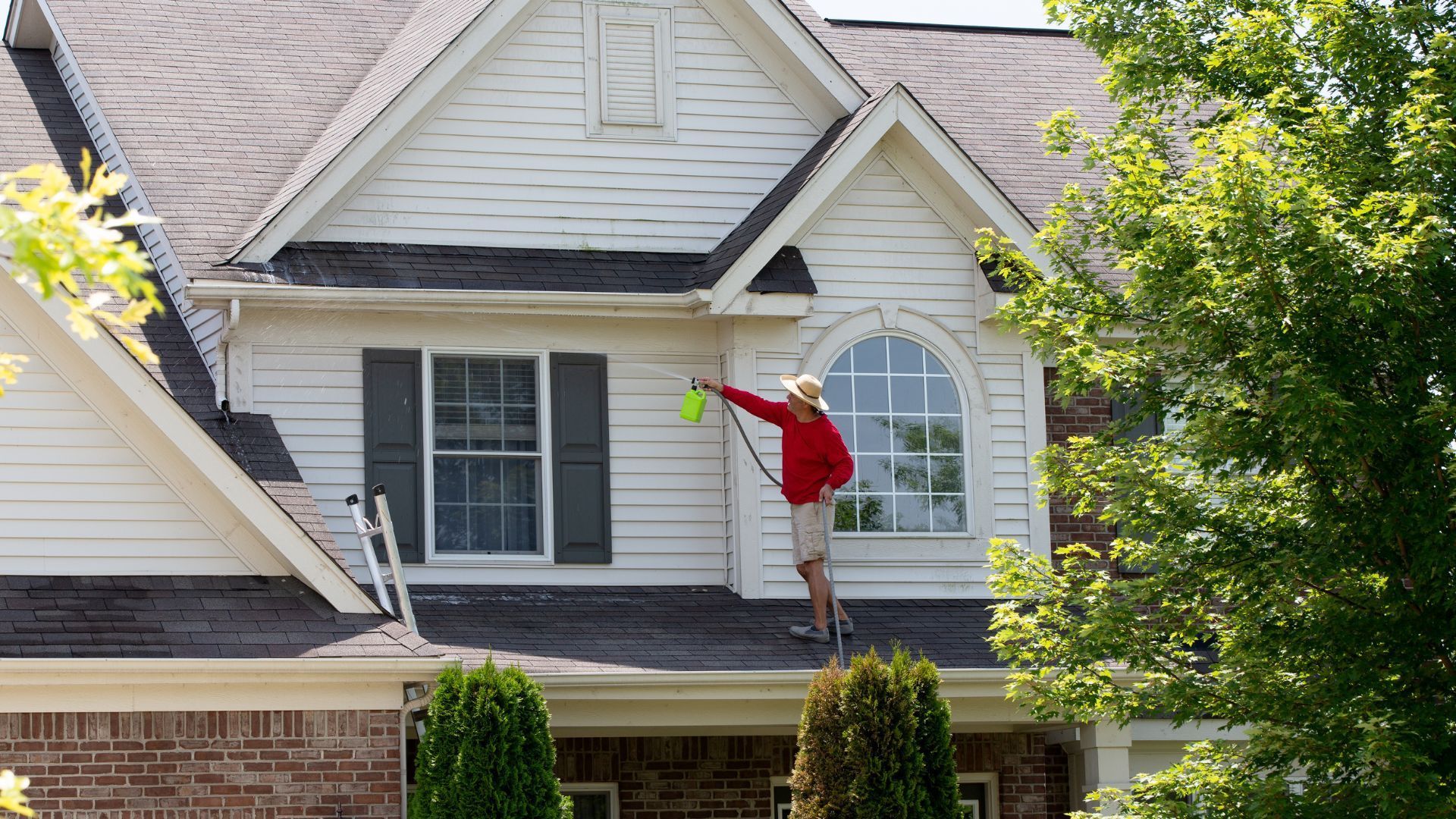 Person in red shirt working on a house roof beside a leafy tree