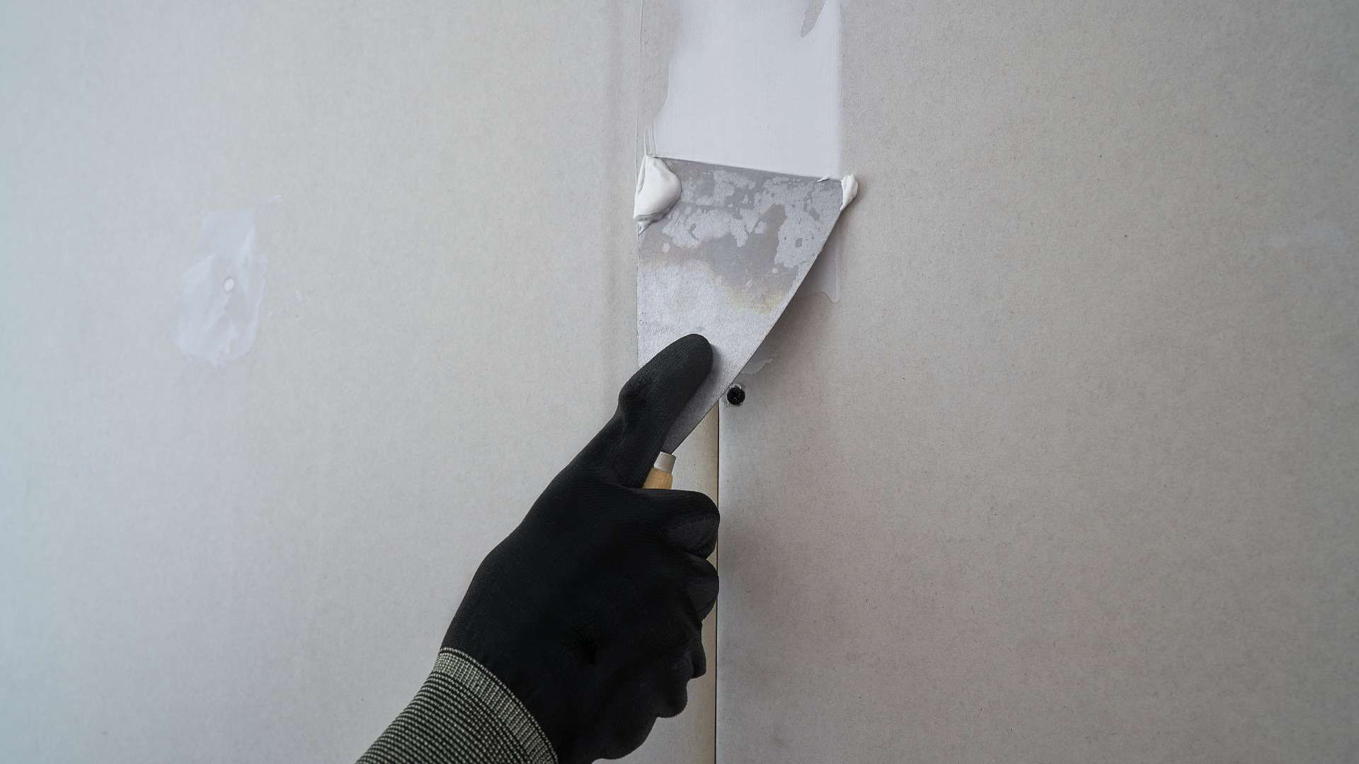 Gloved hand scraping peeling paint from a wall seam with a putty knife