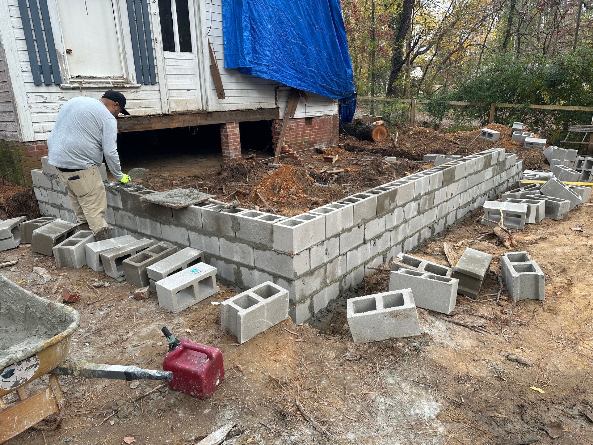 Worker building a gray cinder block retaining wall beside a house with a blue tarp-covered porch.