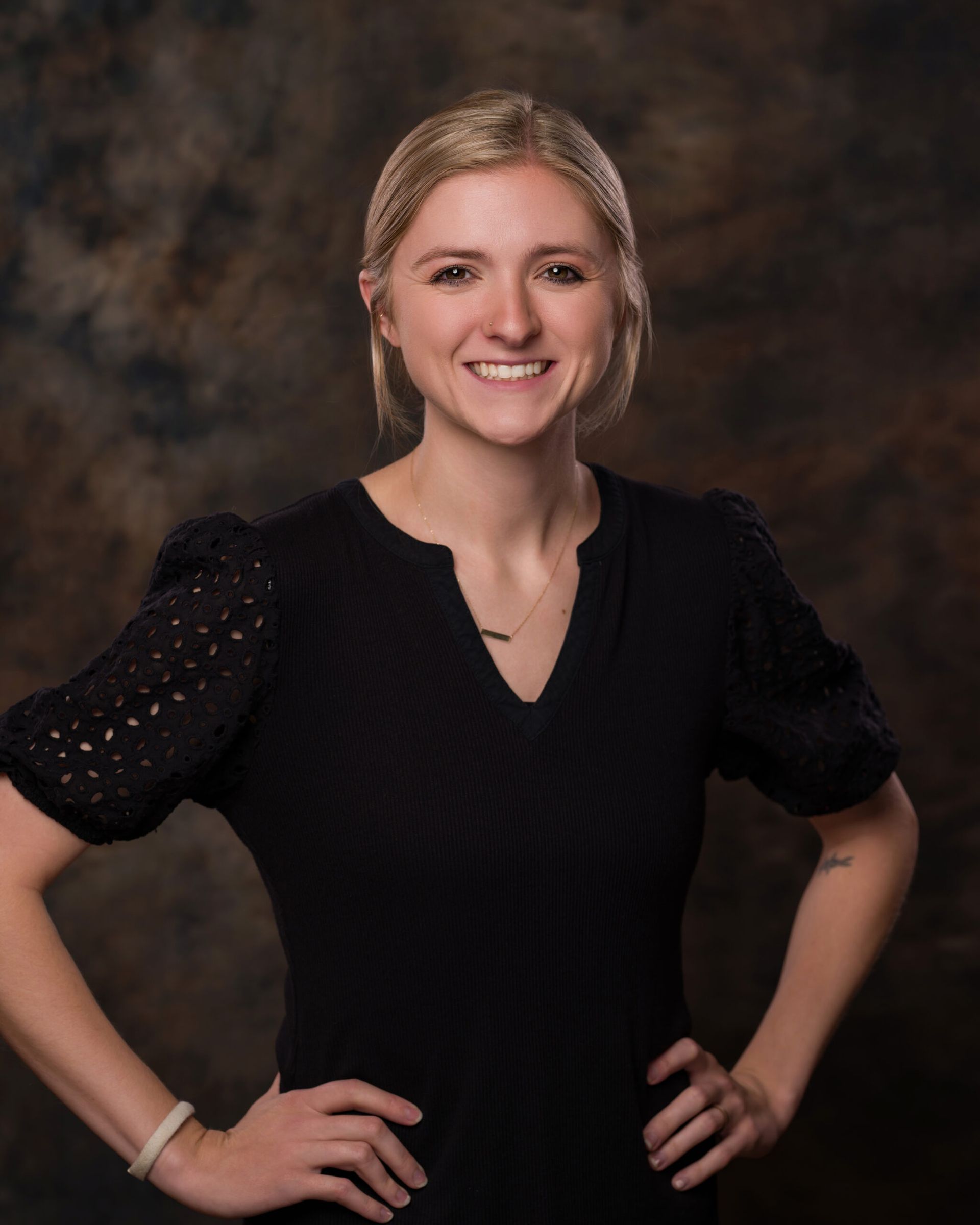 Smiling woman in a black dress with hands on hips against a dark studio backdrop
