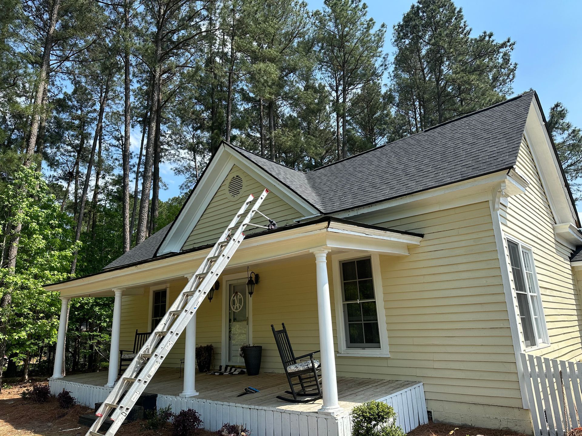 Small beige house with porch, ladder, and dark roof in a wooded yard