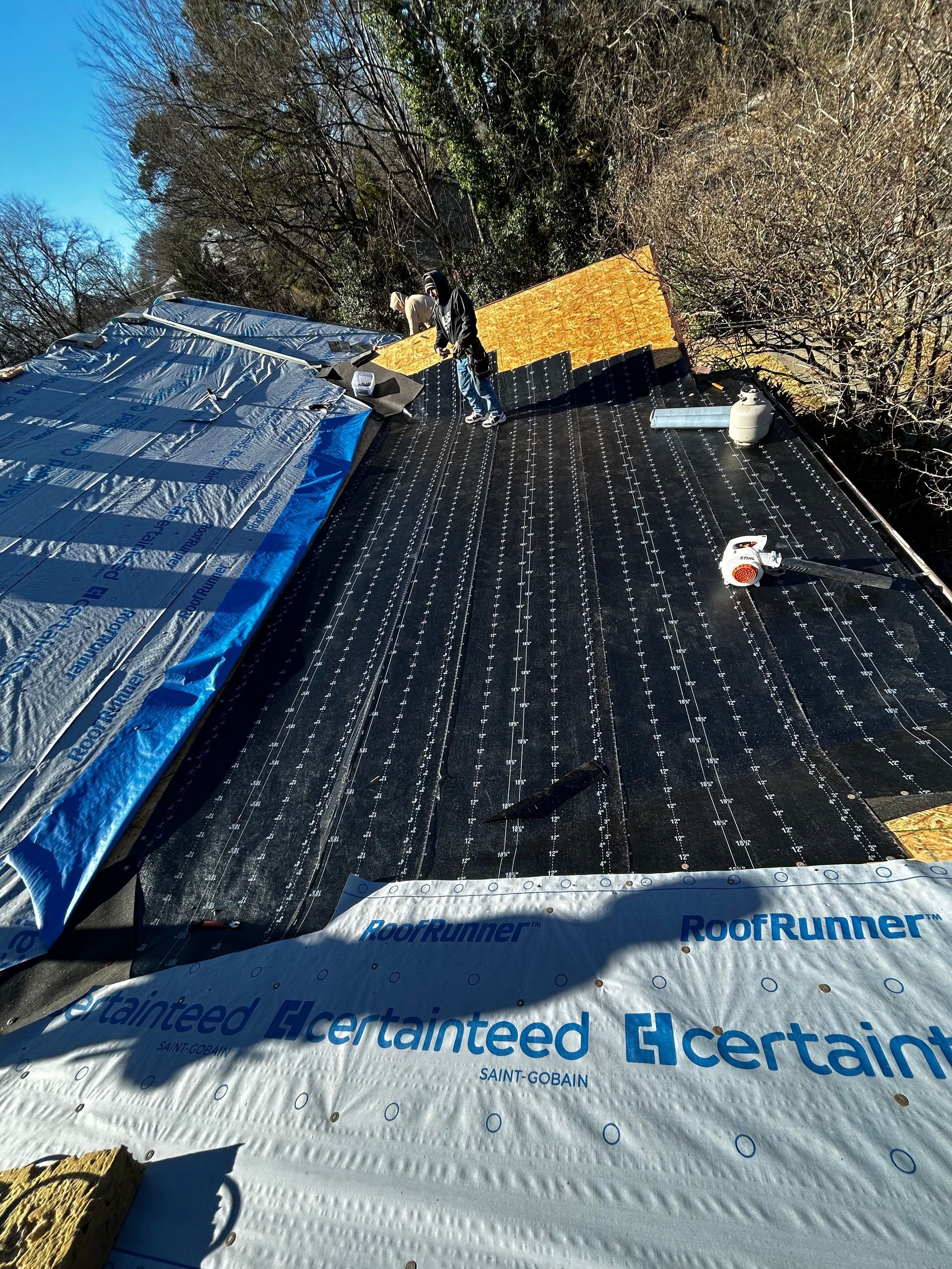 Roof under construction with blue tarp, black underlayment, and yellow roofing materials under trees