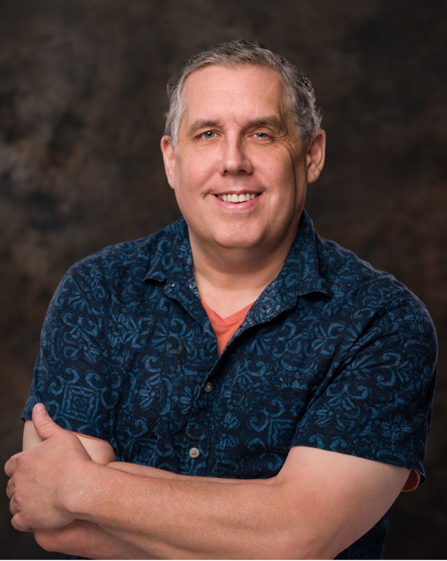Studio portrait of a smiling man in a blue patterned shirt with arms crossed against a dark background