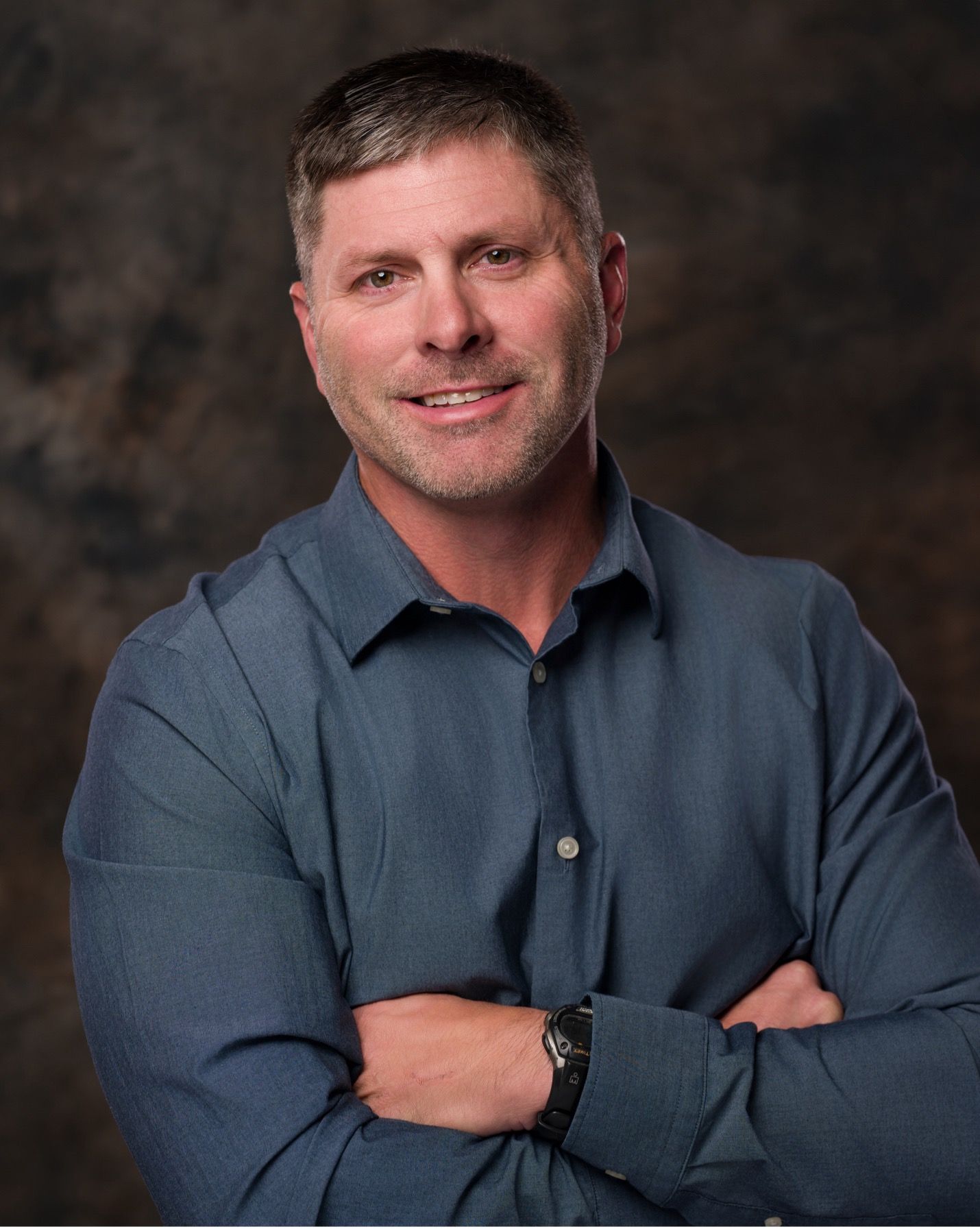 Studio portrait of a smiling man in a blue shirt with arms crossed against a dark background