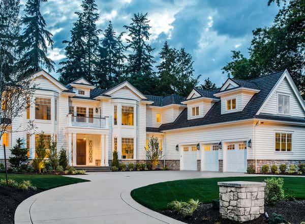 Large white mansion with lit windows, multiple gables, and a curved driveway at dusk