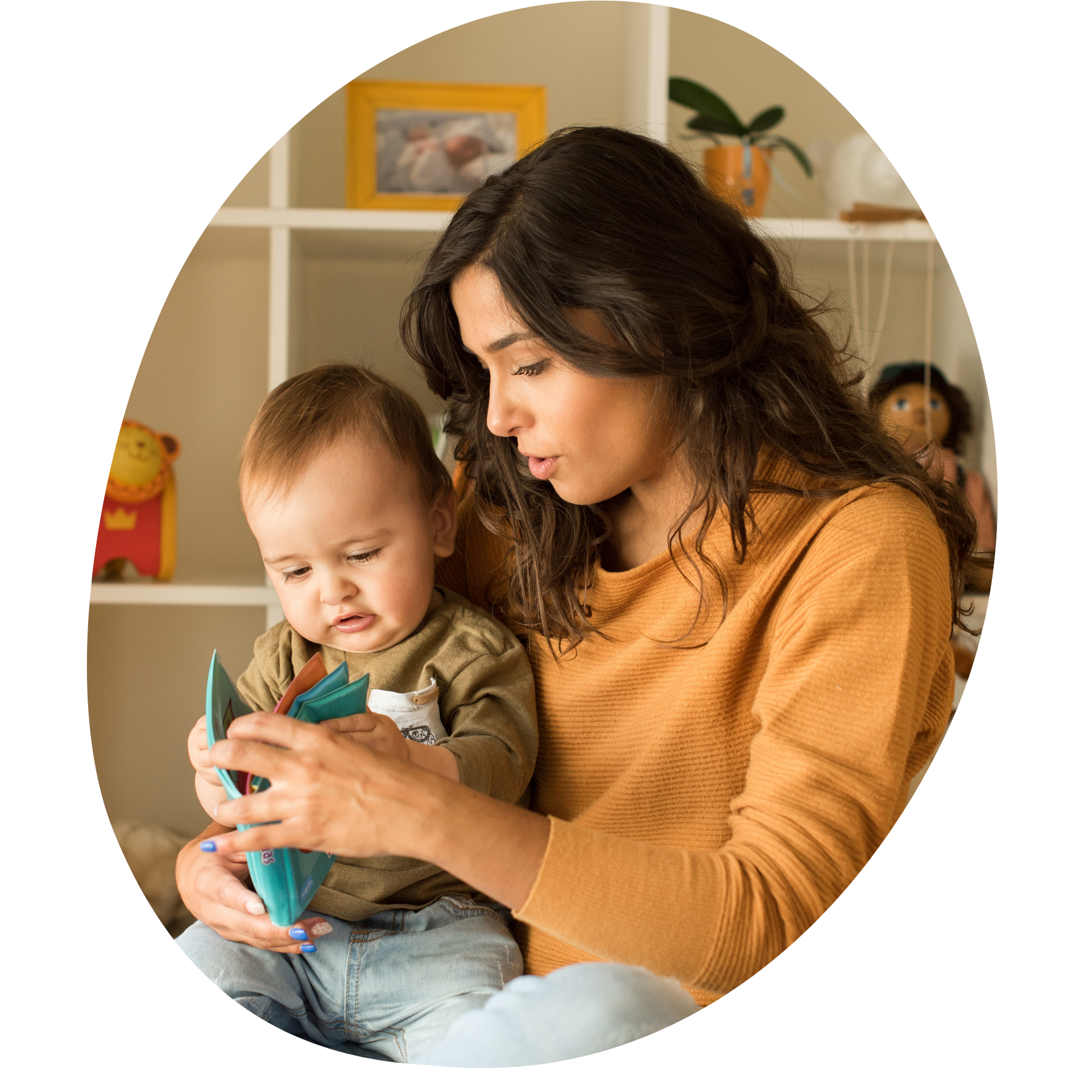 A person in a mustard sweater reads a book with a small child in their lap in a room with shelves and toys.