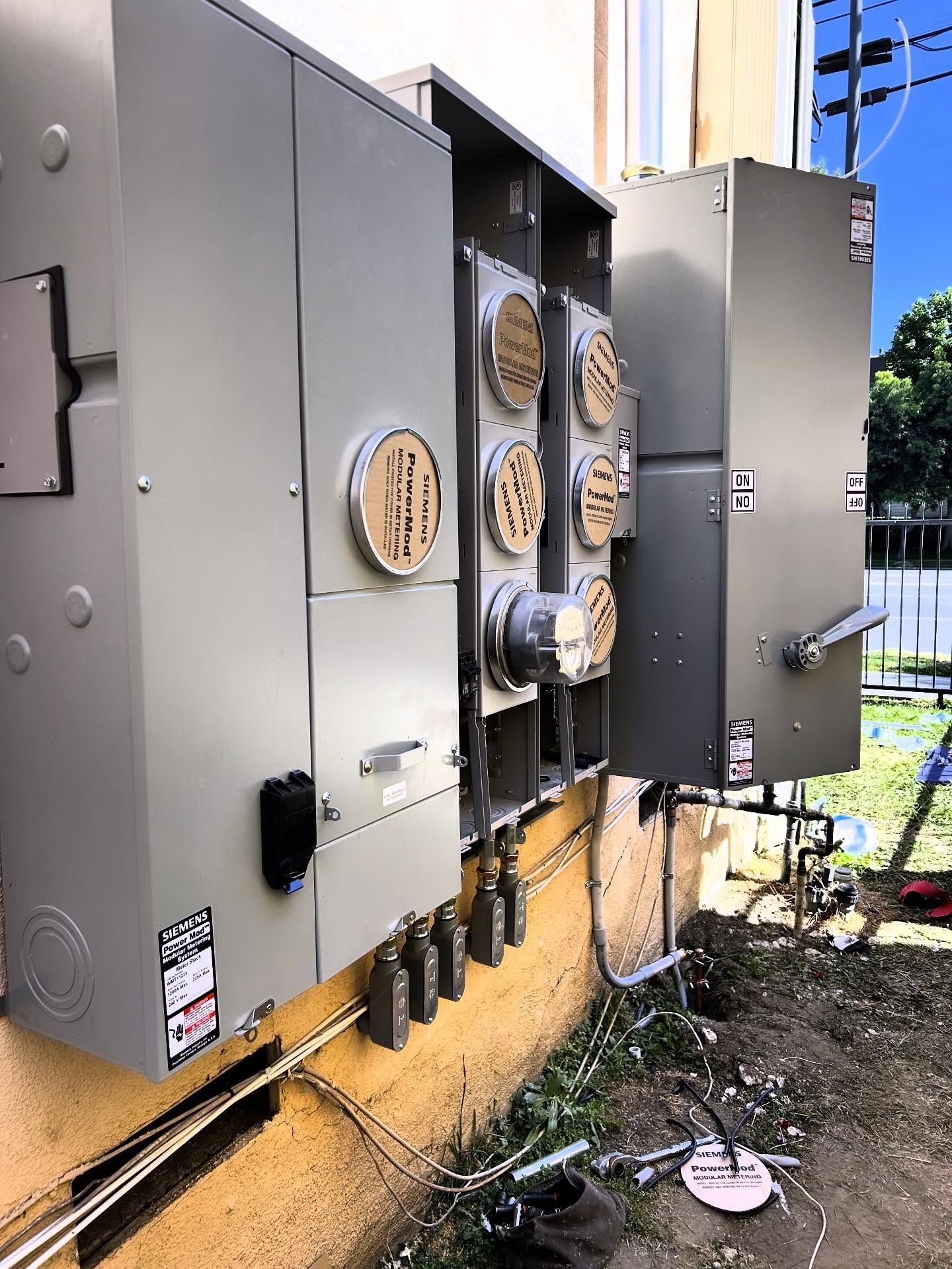 Outdoor electrical service panels and utility boxes mounted on a wall beside a sidewalk and plants.