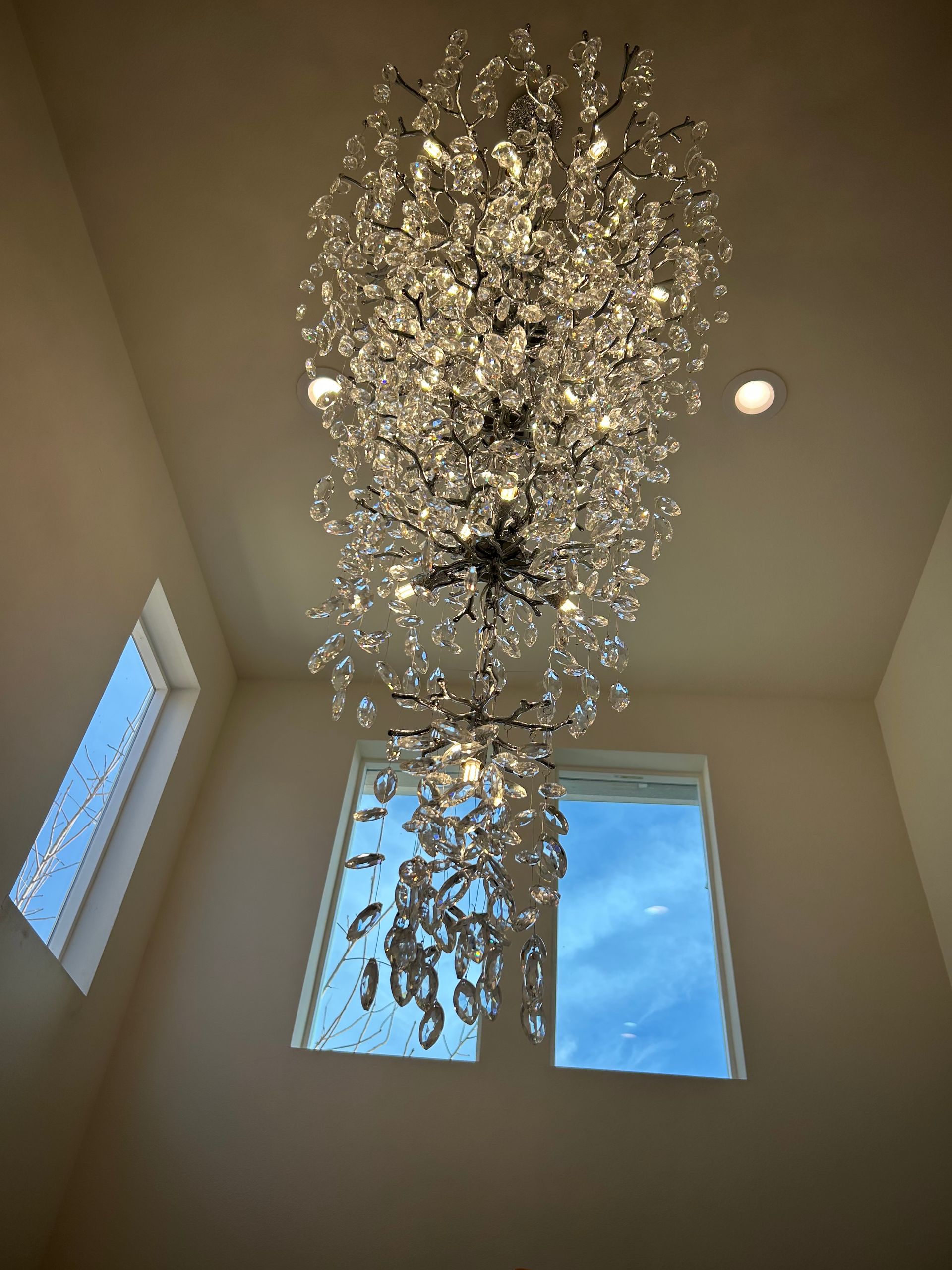 A tall crystal chandelier hangs from a high ceiling beside two windows with daylight.
