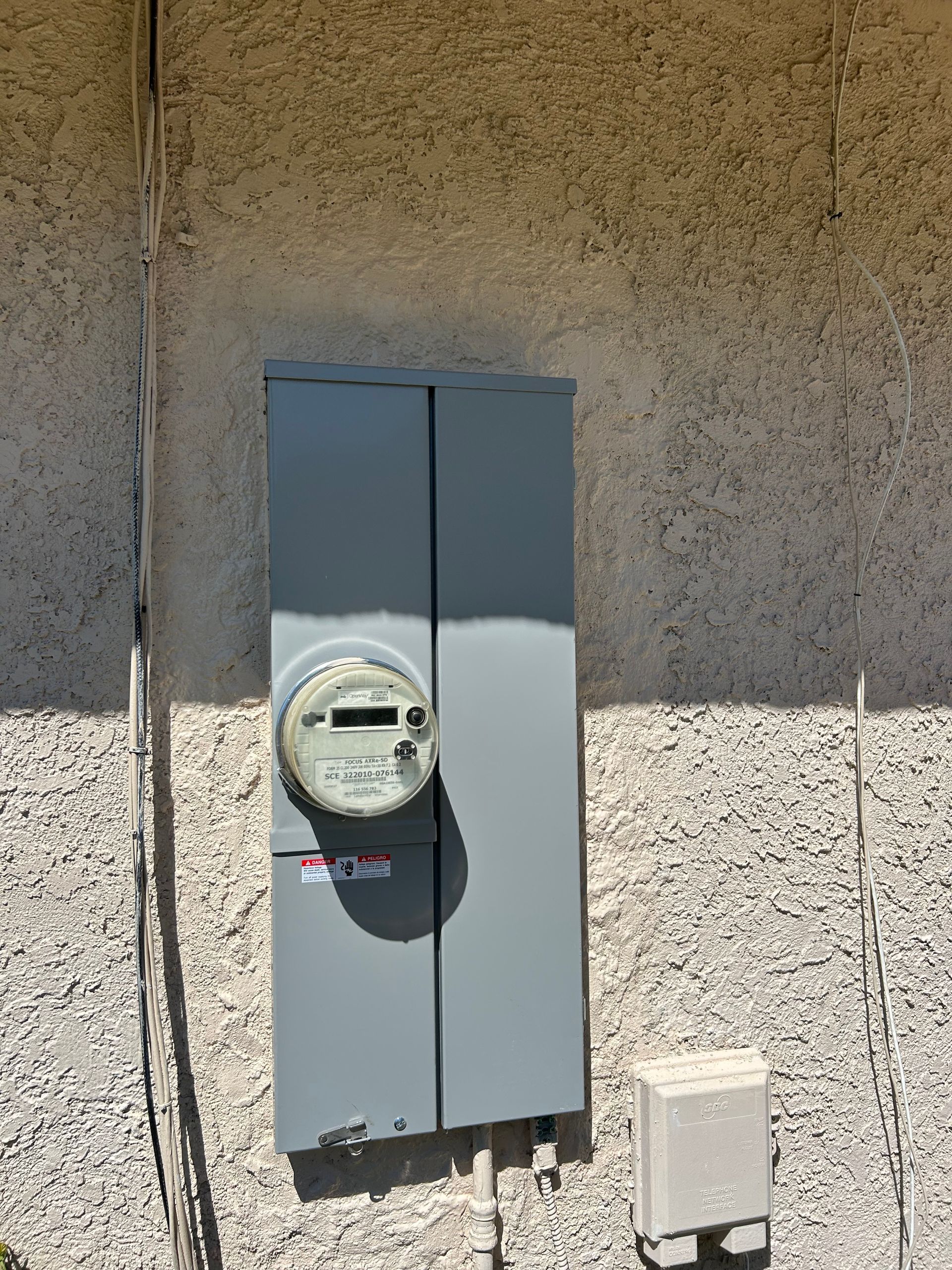 Gray electrical meter box mounted on a stucco wall, with conduit and a small utility box below.