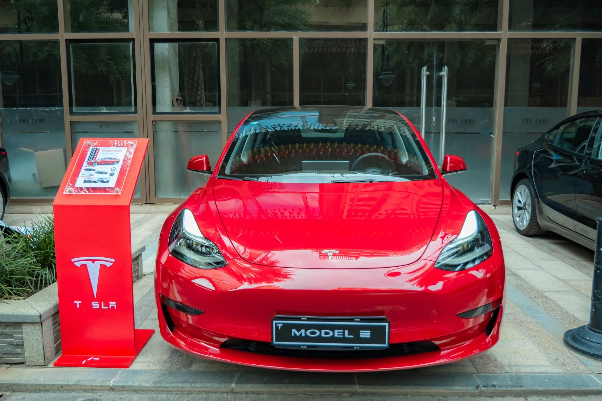 Red Tesla Model 3 displayed indoors on a showroom floor beside a charging stand.