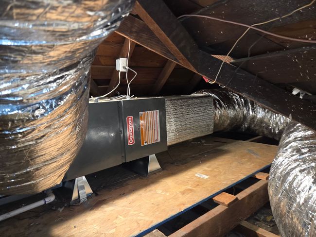 Technician in blue coveralls testing an open furnace cabinet with a multimeter inside a home.