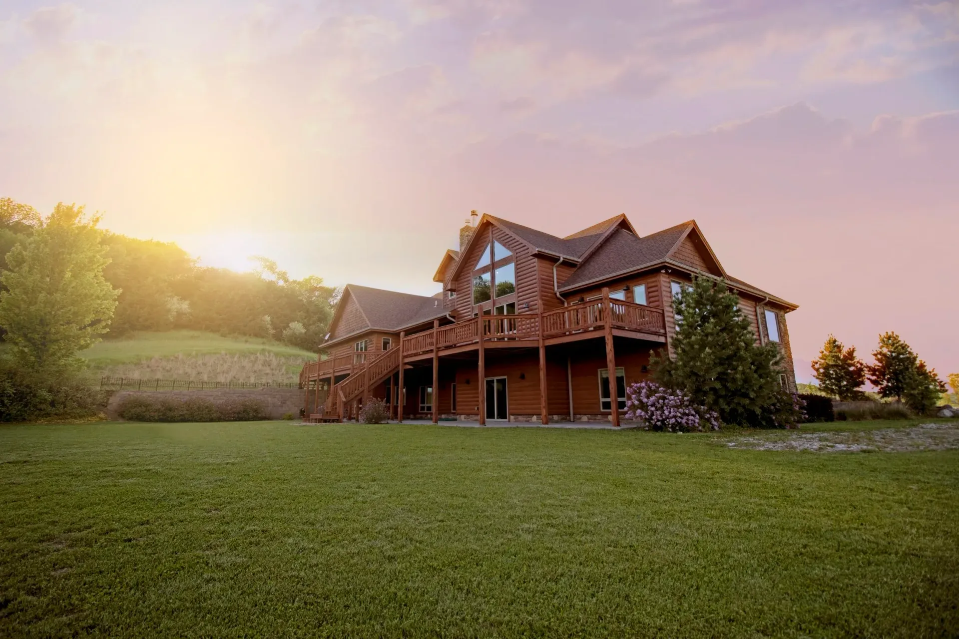 Large wooden house on a grassy lawn at sunset, with trees and a warm pink sky.