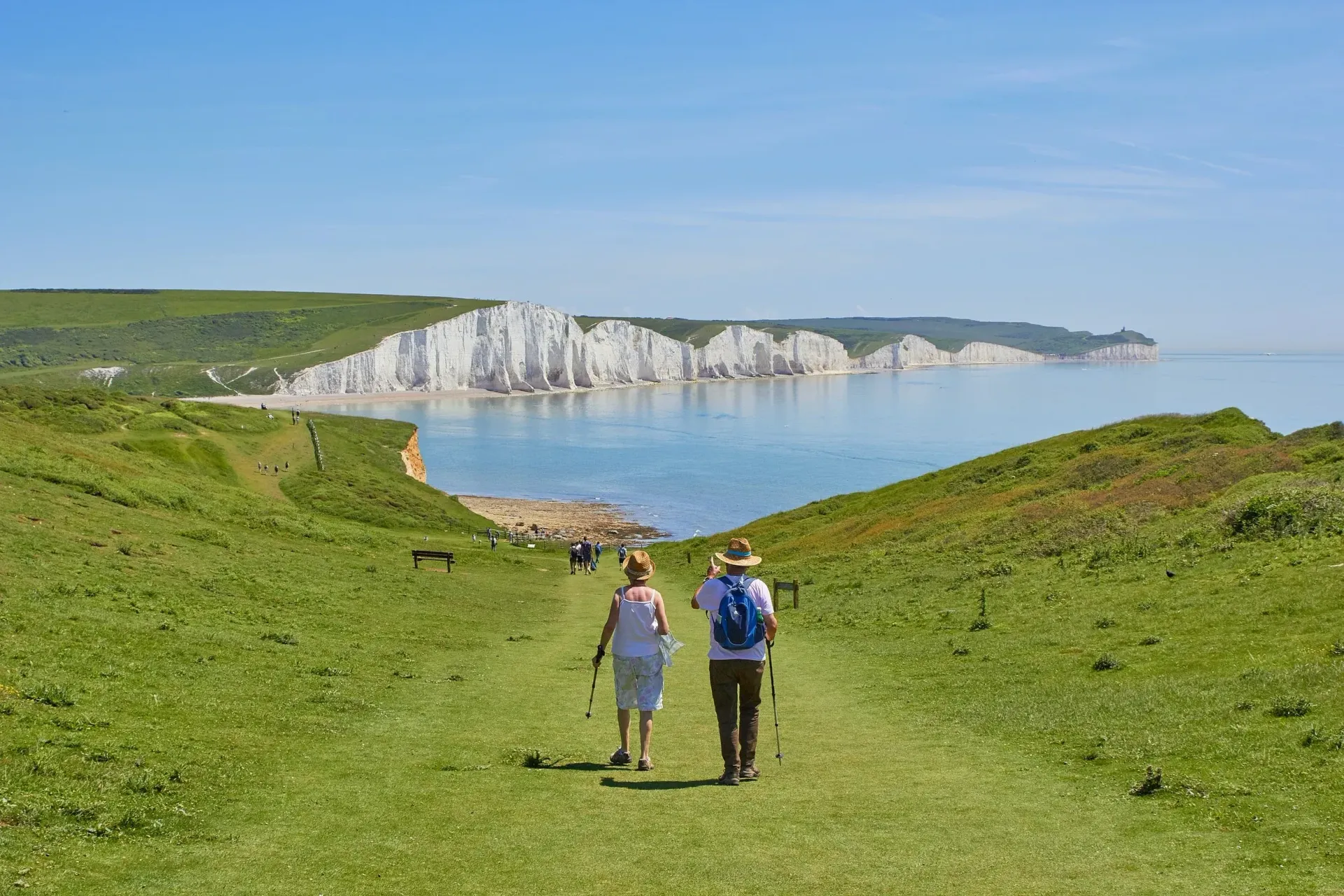 Two hikers walking down a grassy path toward a white chalk cliff and calm blue sea under a clear sky