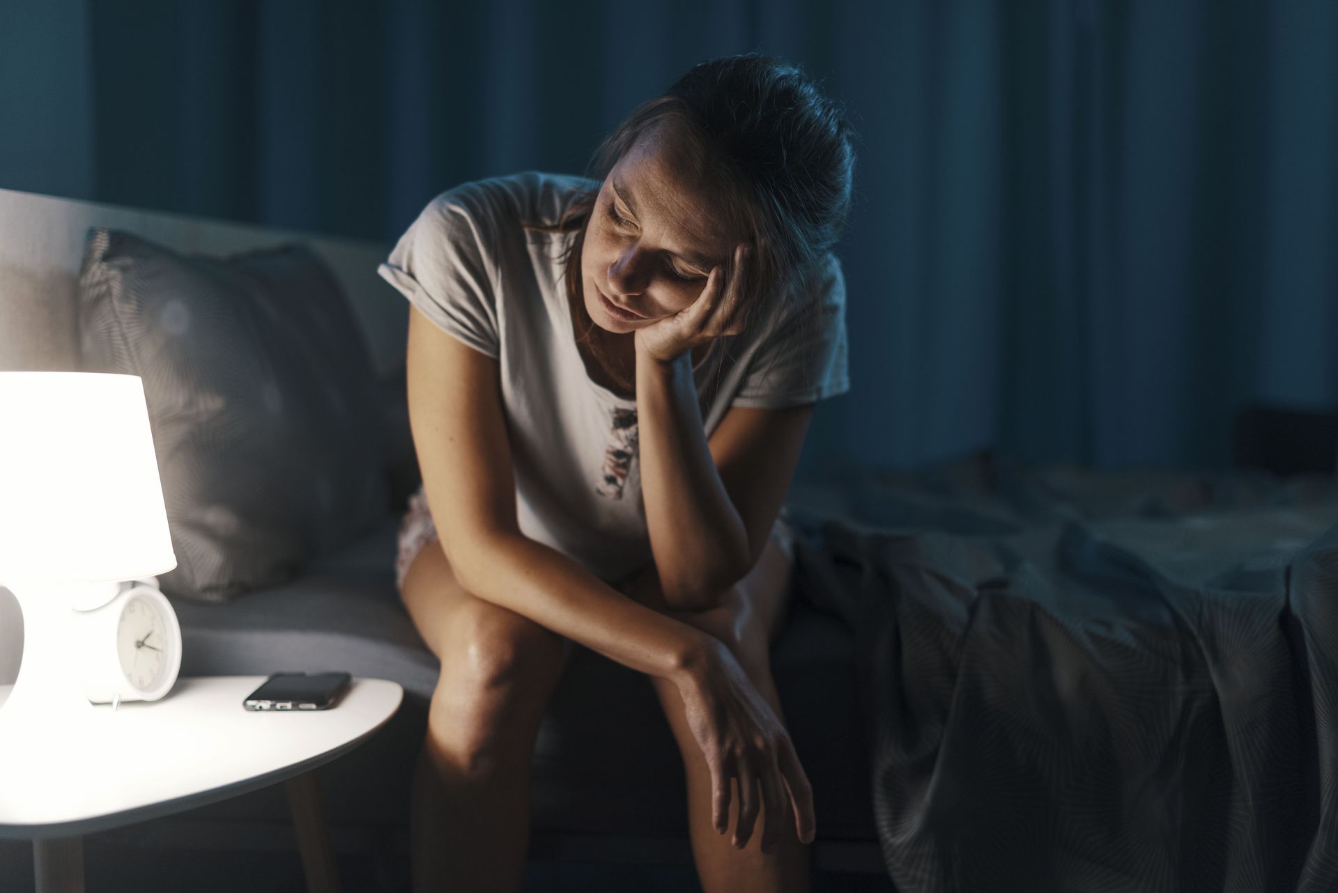 Uma mulher está sentada na cama com a cabeça entre as mãos.