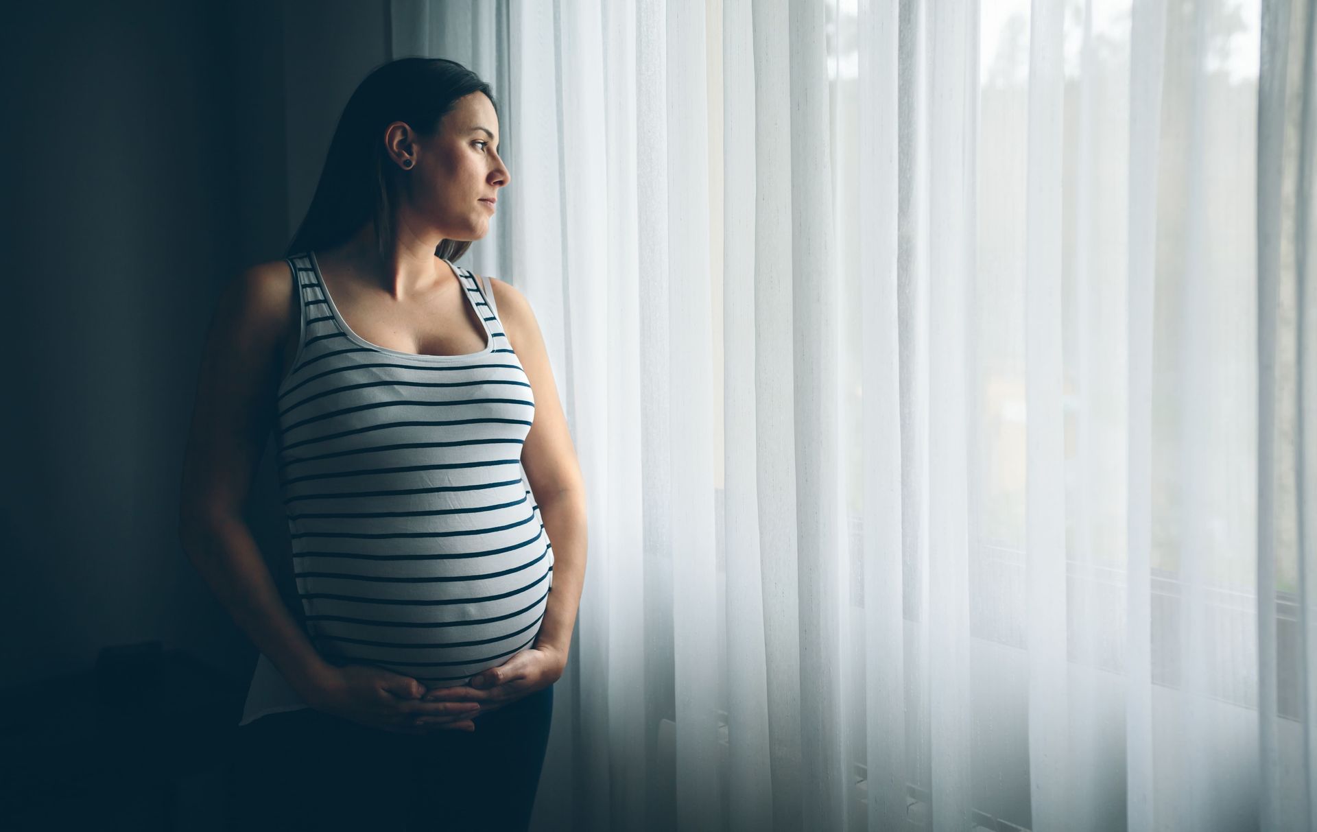 Uma mulher grávida está parada em frente a uma janela segurando sua barriga.