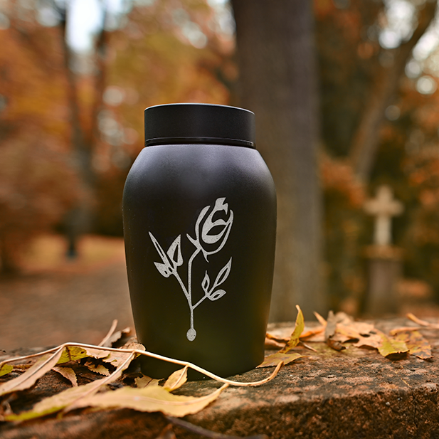 Black urn with a rose engraving, set outdoors with autumn leaves and a blurred cemetery background.