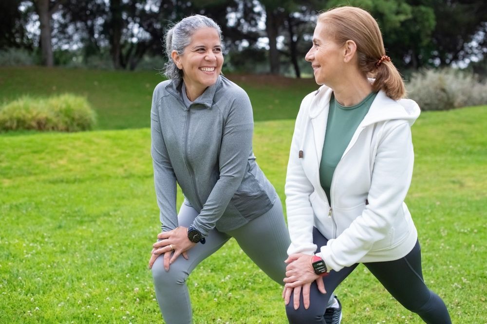 Two women stretching in a grassy park, smiling and facing each other.