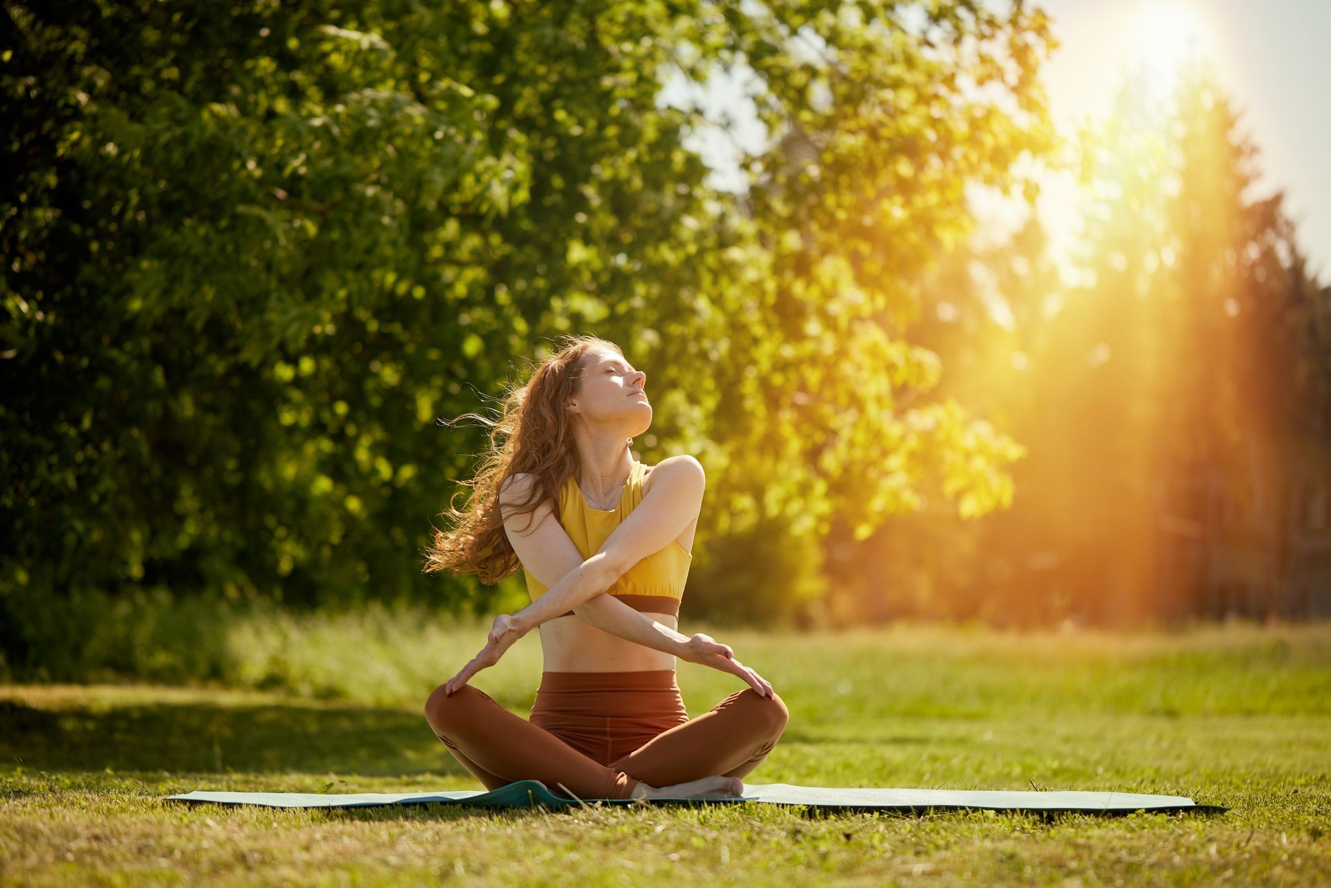 Woman sitting cross-legged on a yoga mat in a sunny park, eyes closed, stretching in golden light
