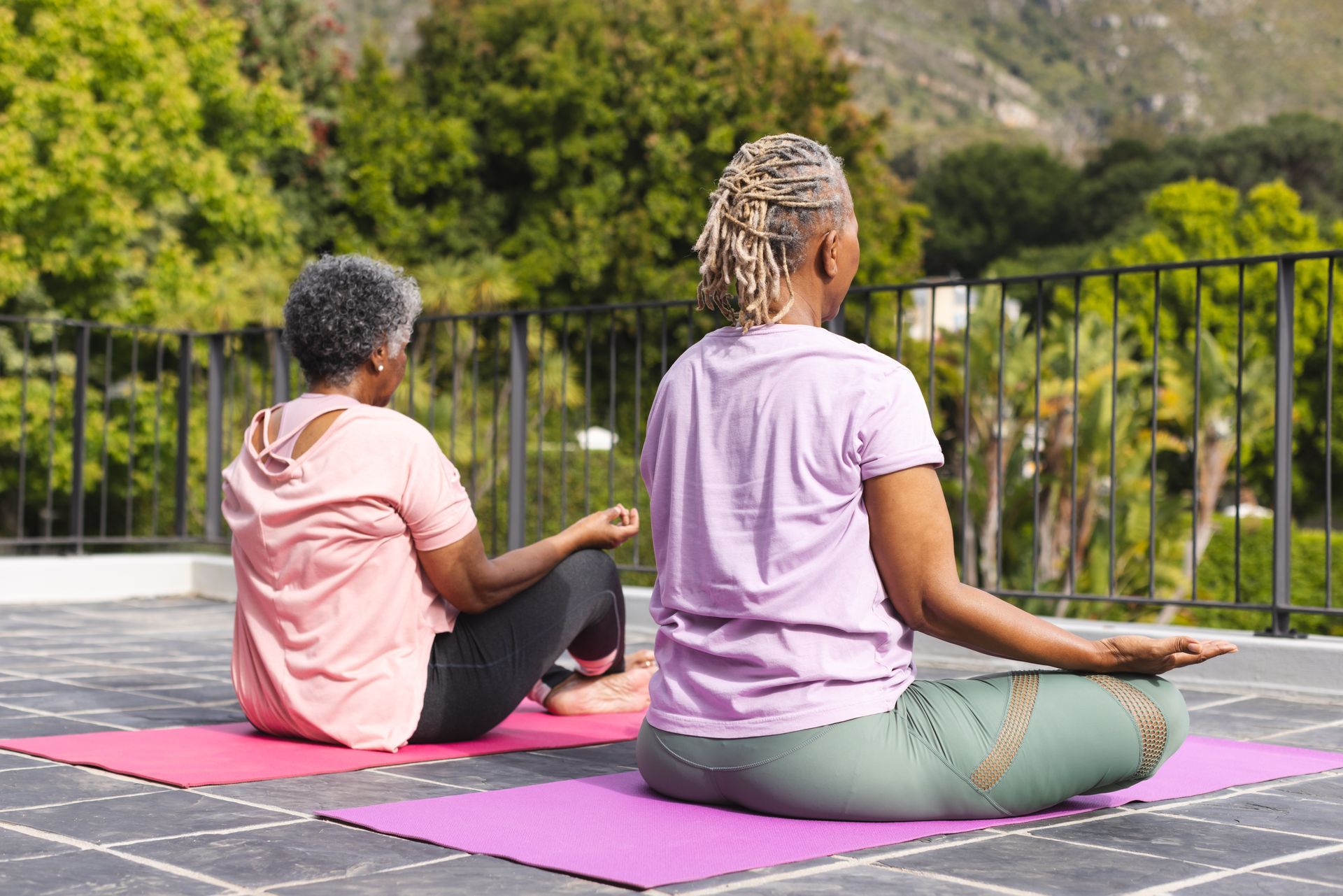 Two people sitting on yoga mats outdoors, facing a green hillside.