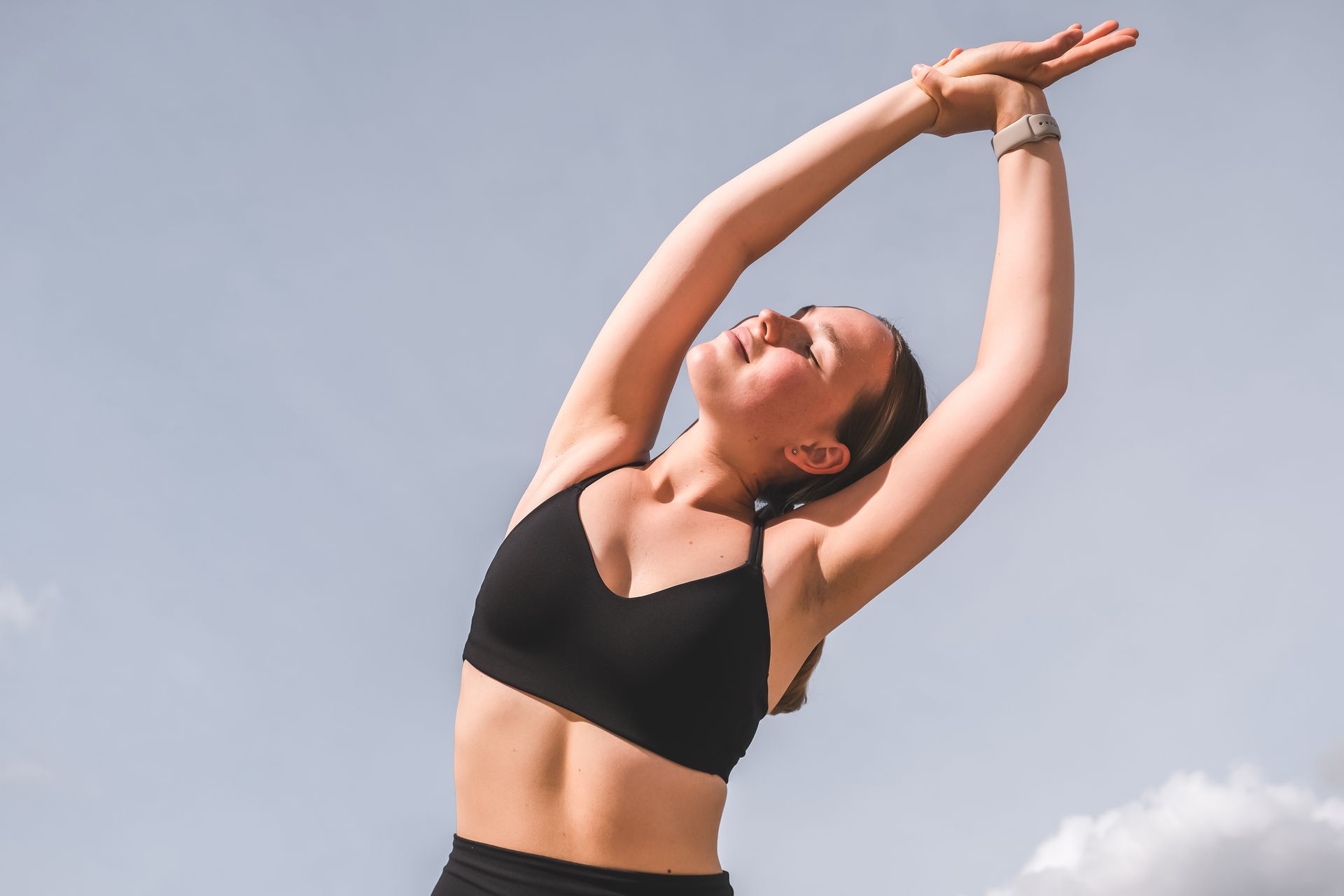Woman stretching outdoors against a blue sky, wearing a black sports bra.