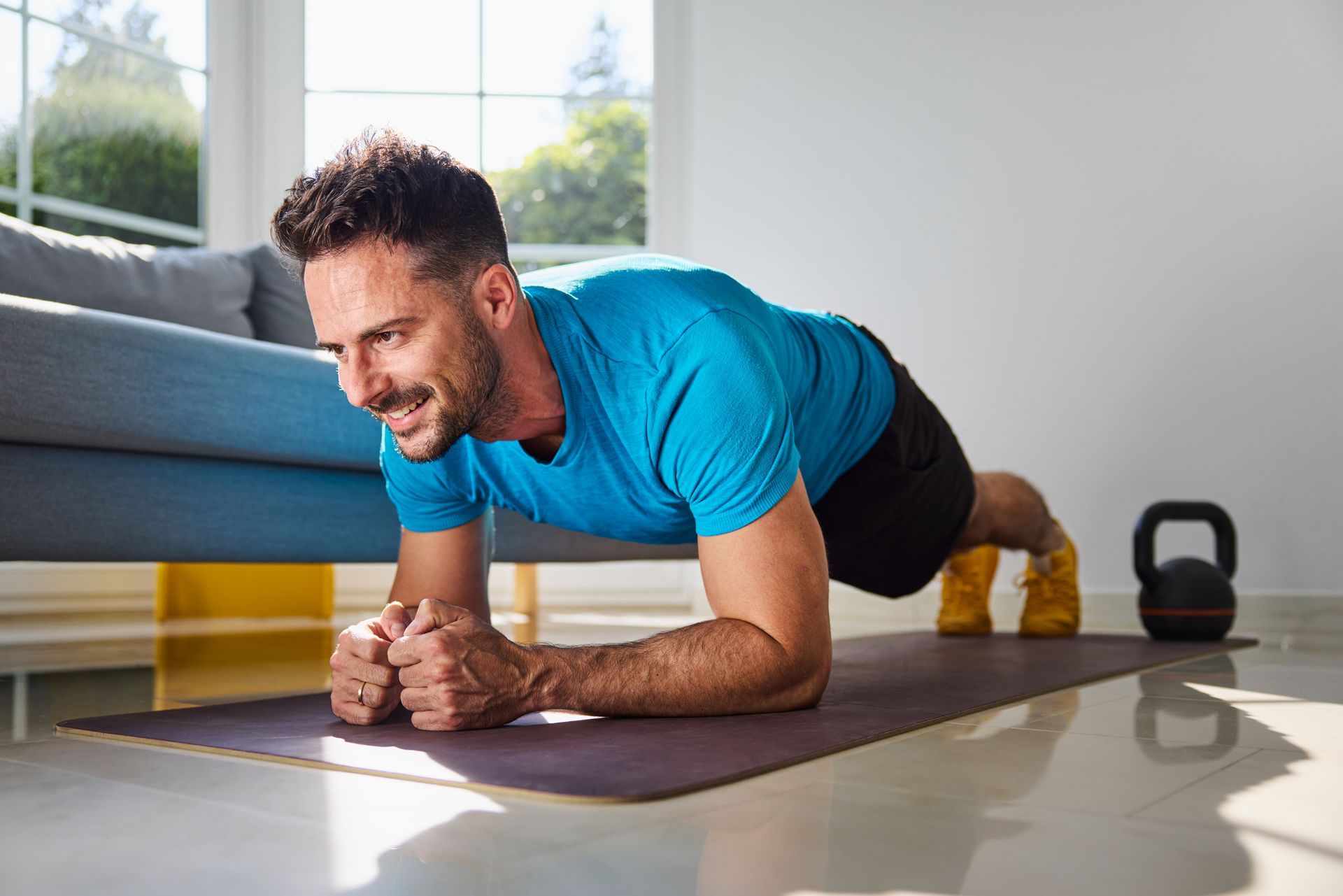 Man planks on a yoga mat in a bright room, smiling beside a gray sofa and kettlebell.