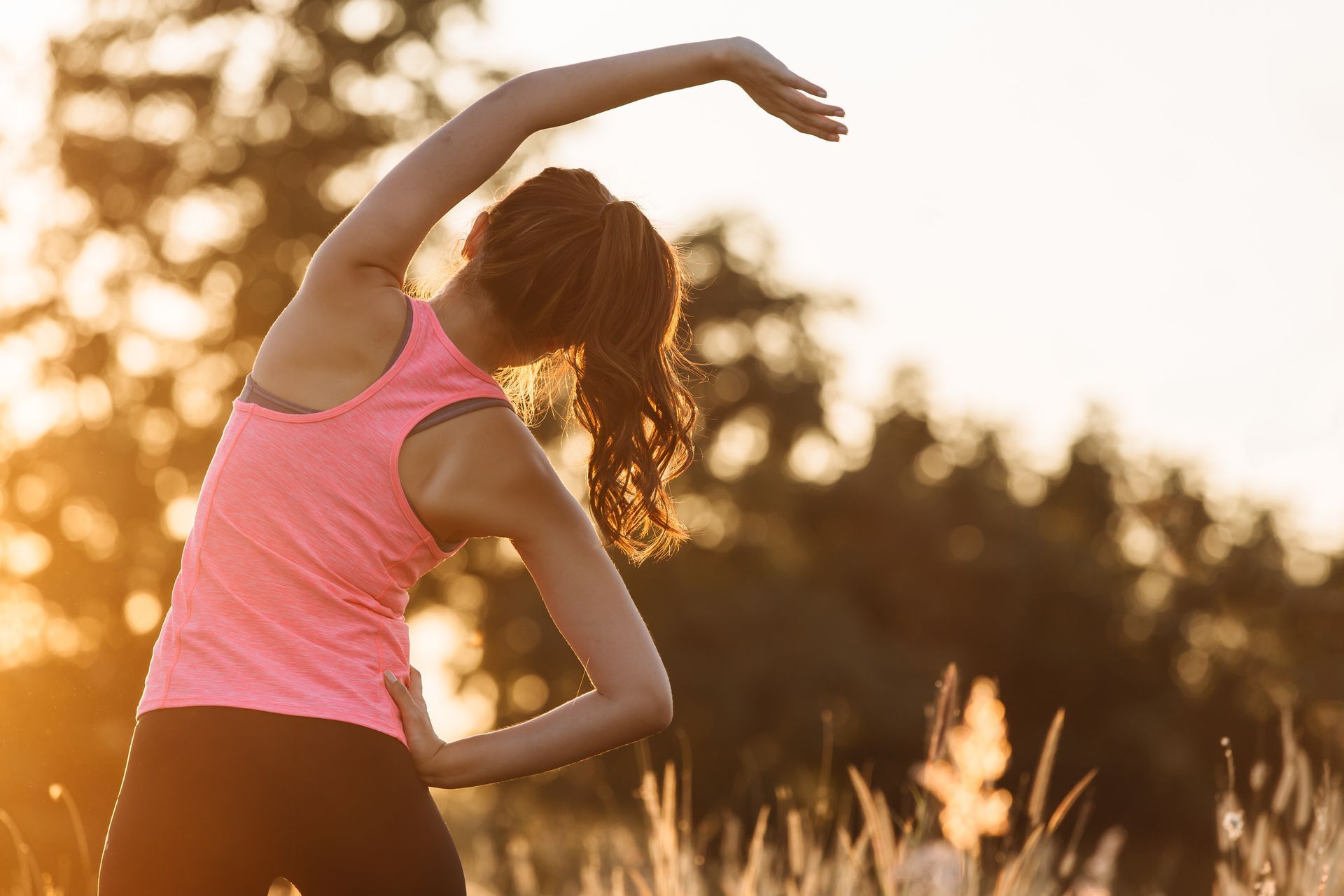 Woman stretching outdoors in a pink tank top at sunset.