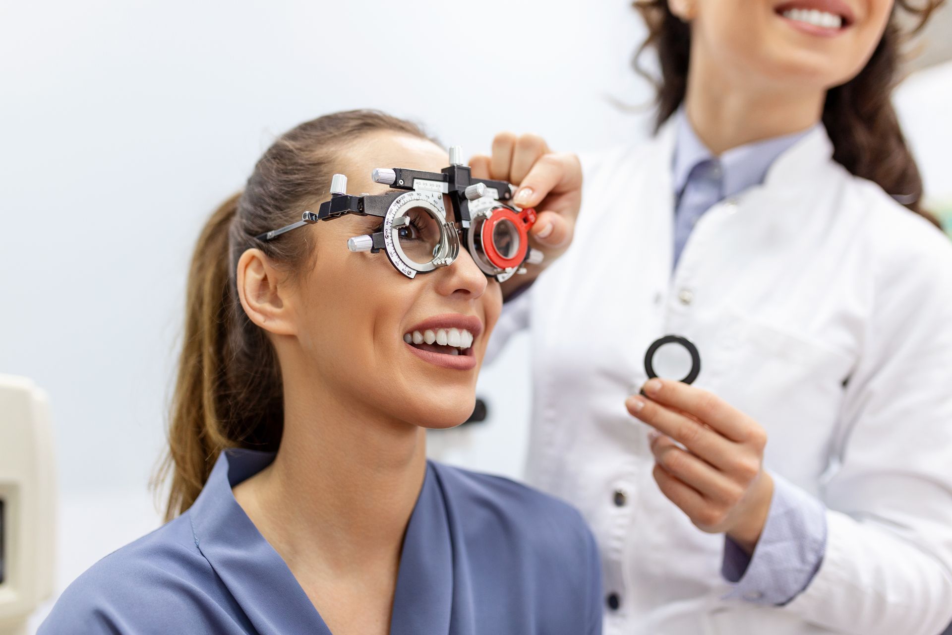Woman in an eye exam, wearing a phoropter while an optometrist adjusts a lens at her side