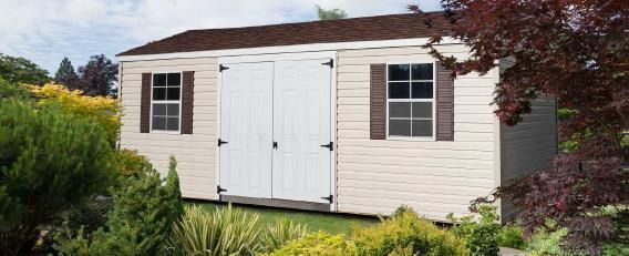 White garden shed with double doors and windows, surrounded by green shrubs and flowers