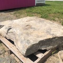 Large flat gray stone slab resting on a wooden pallet outdoors beside grass and a building