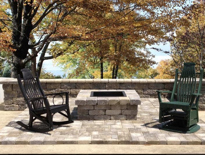 Two rocking chairs facing a stone fire pit on a brick patio under autumn trees