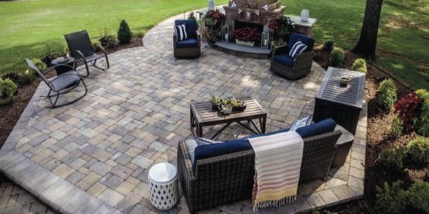 Outdoor patio with wicker seating, stone pavers, fire pit, and surrounding lawn and trees.