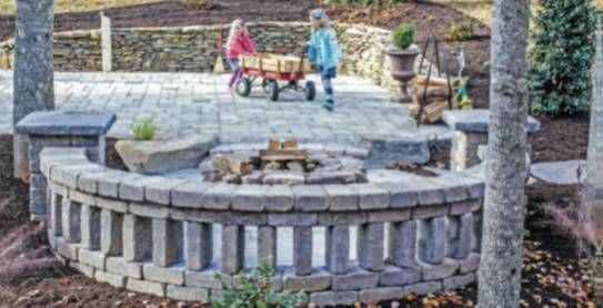 Stone circular fire pit in a landscaped yard, with two people standing near a red wagon in the background.