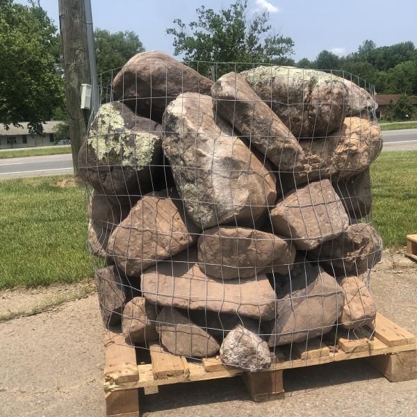 Large decorative rock pile stacked on a wooden pallet, wrapped in wire mesh outdoors near a roadside.