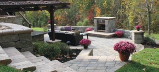 Stone patio with steps, a fire pit, and pink potted flowers in a landscaped backyard.